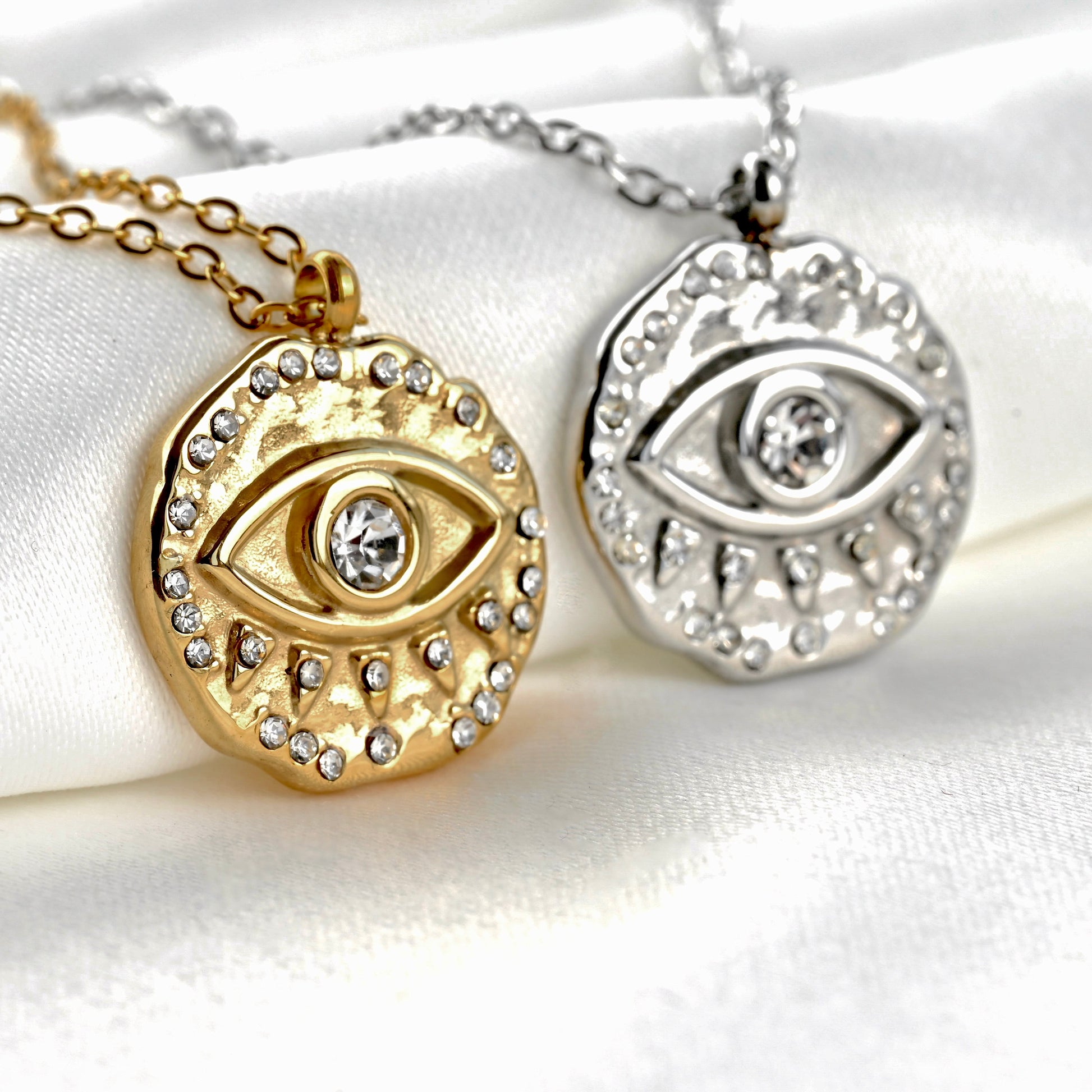 Close-up view of one gold and one silver antique looking coin pendants with embossed evil eye design in the center, on a white fabric background.