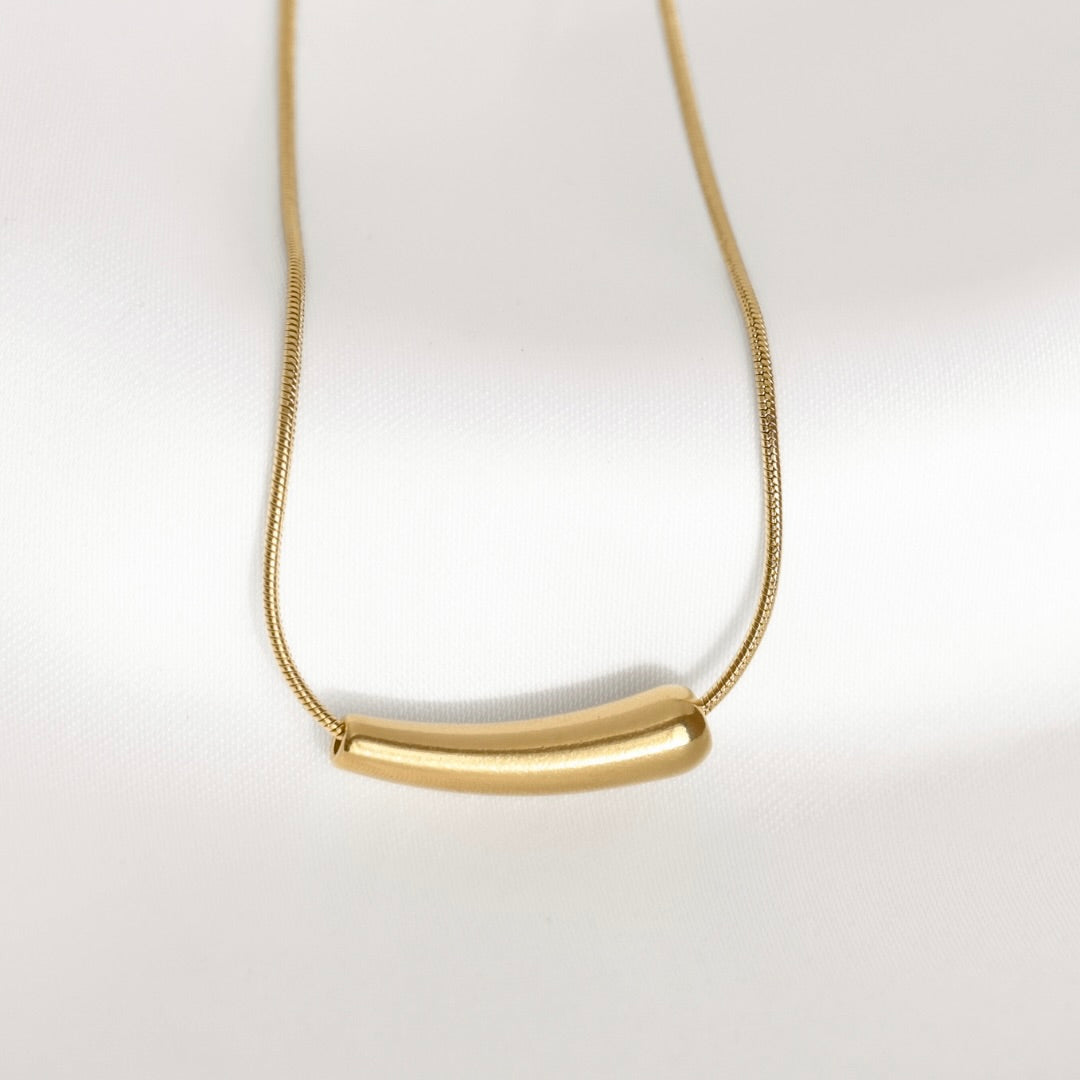 A gold necklace with a curved bar pendant on a delicate chain, photographed against a clean white background.