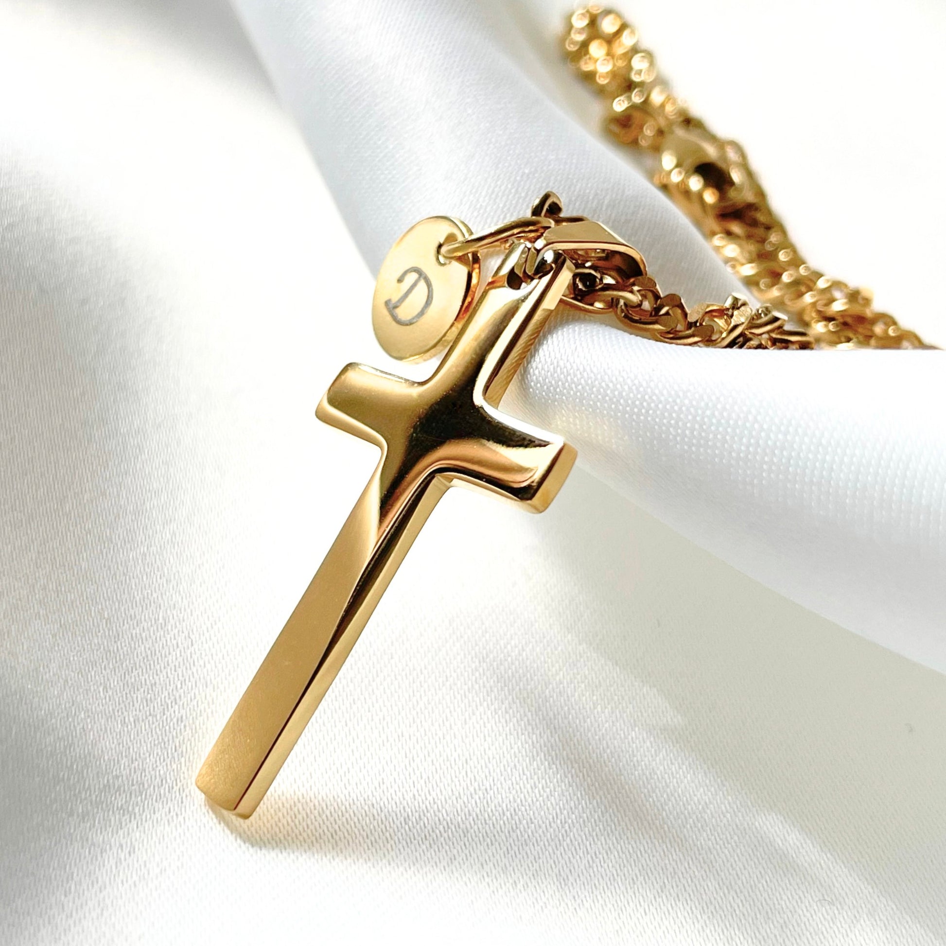 Gold cross necklace with a small round initial charm on a white fabric background