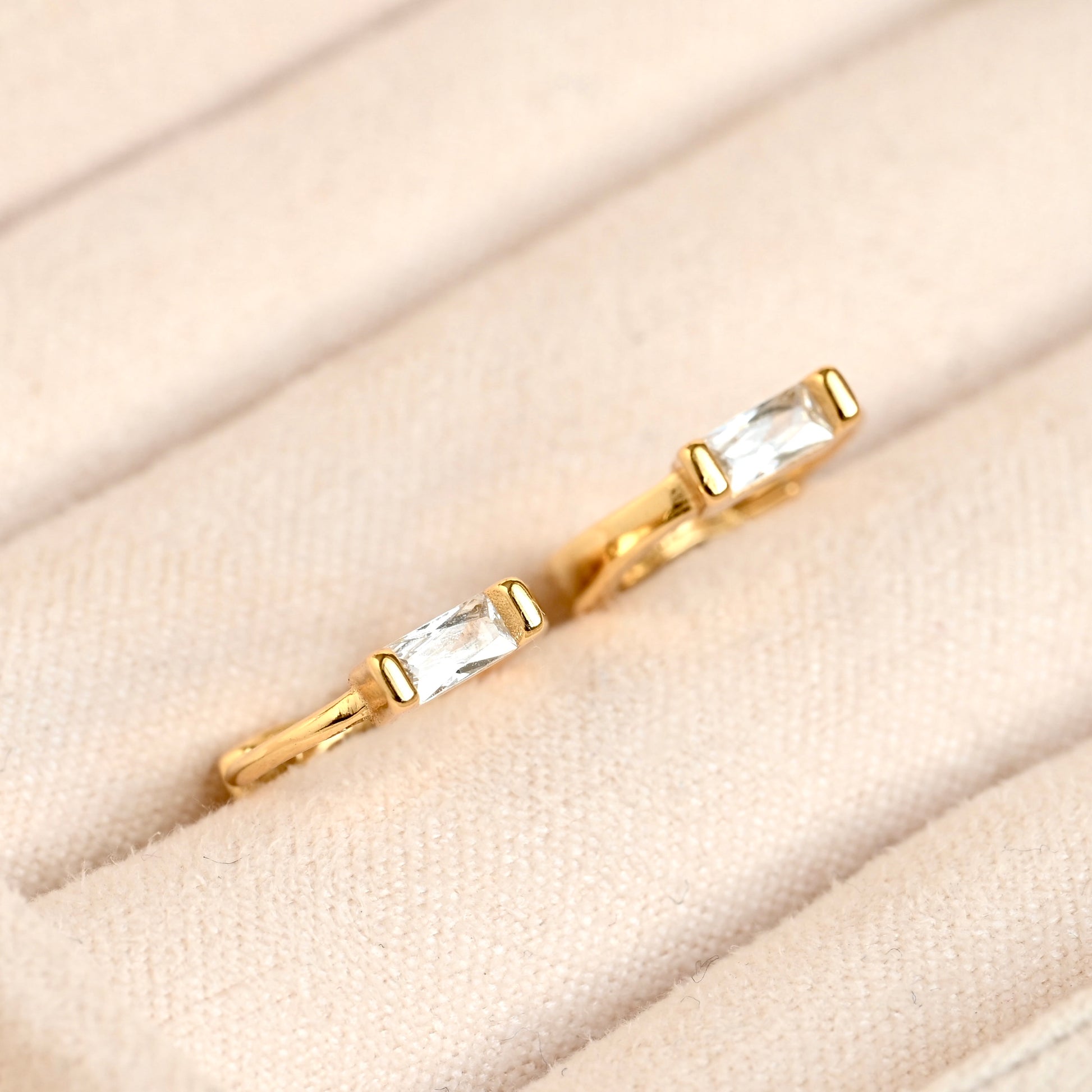 A pair of gold rings with rectangular clear gemstones, displayed on a beige textured ring box.
