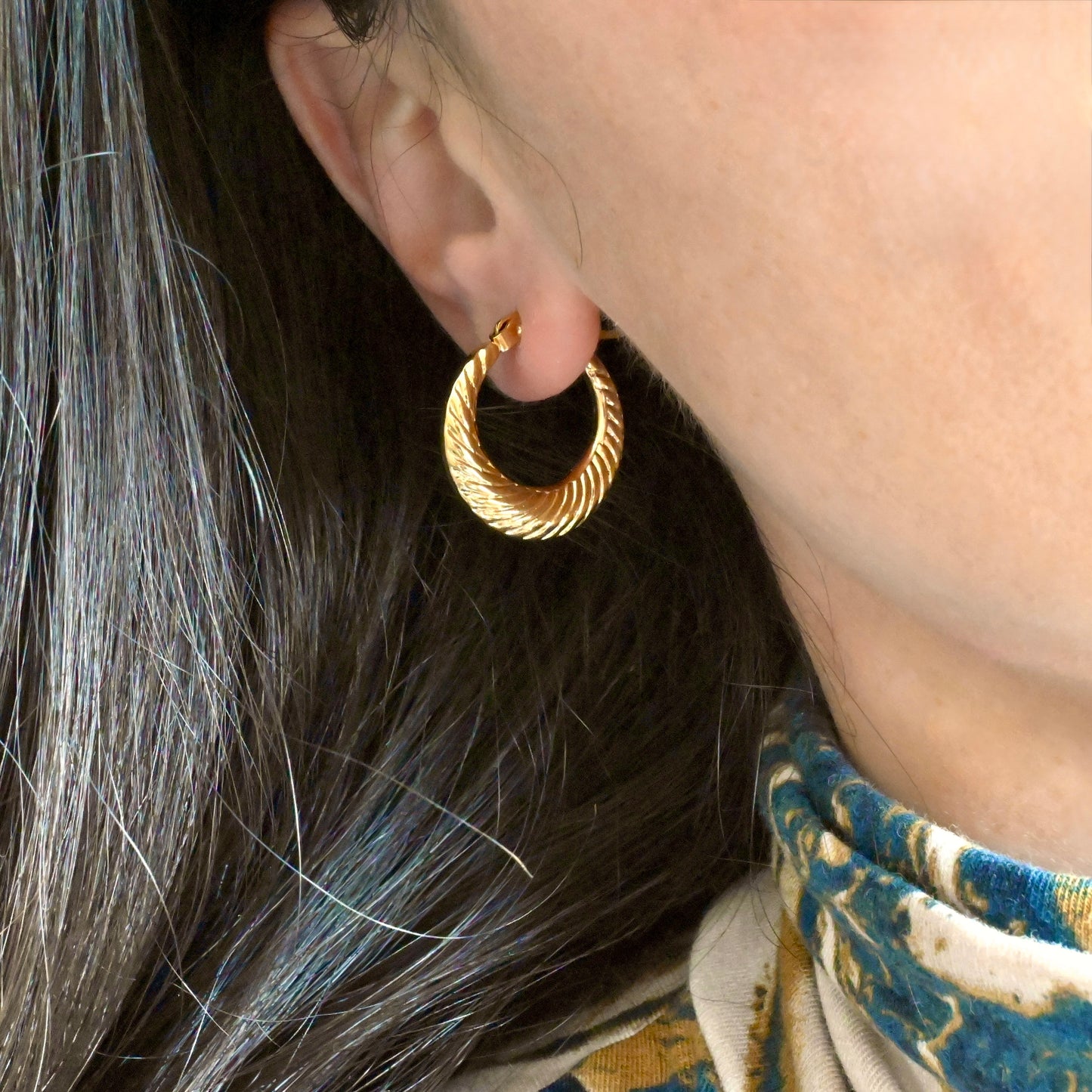 Close-up of a person wearing a small ribbed gold hoop earrings with hair and fabric in the background.