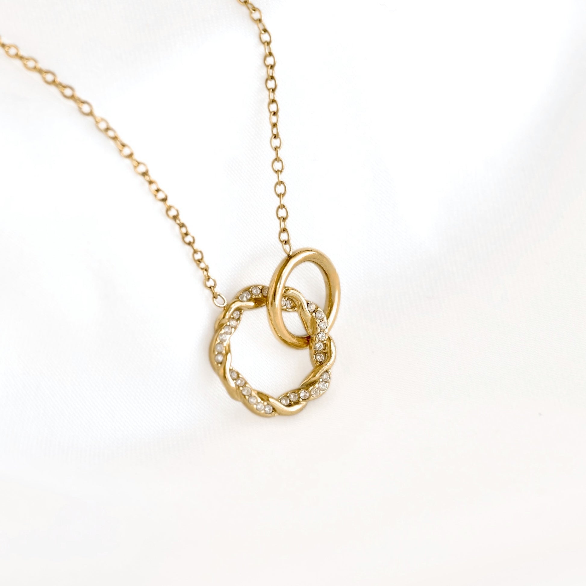 A gold chain necklace with a pendant featuring two interlocking rings, one adorned with small clear stones, photographed on a white background.
