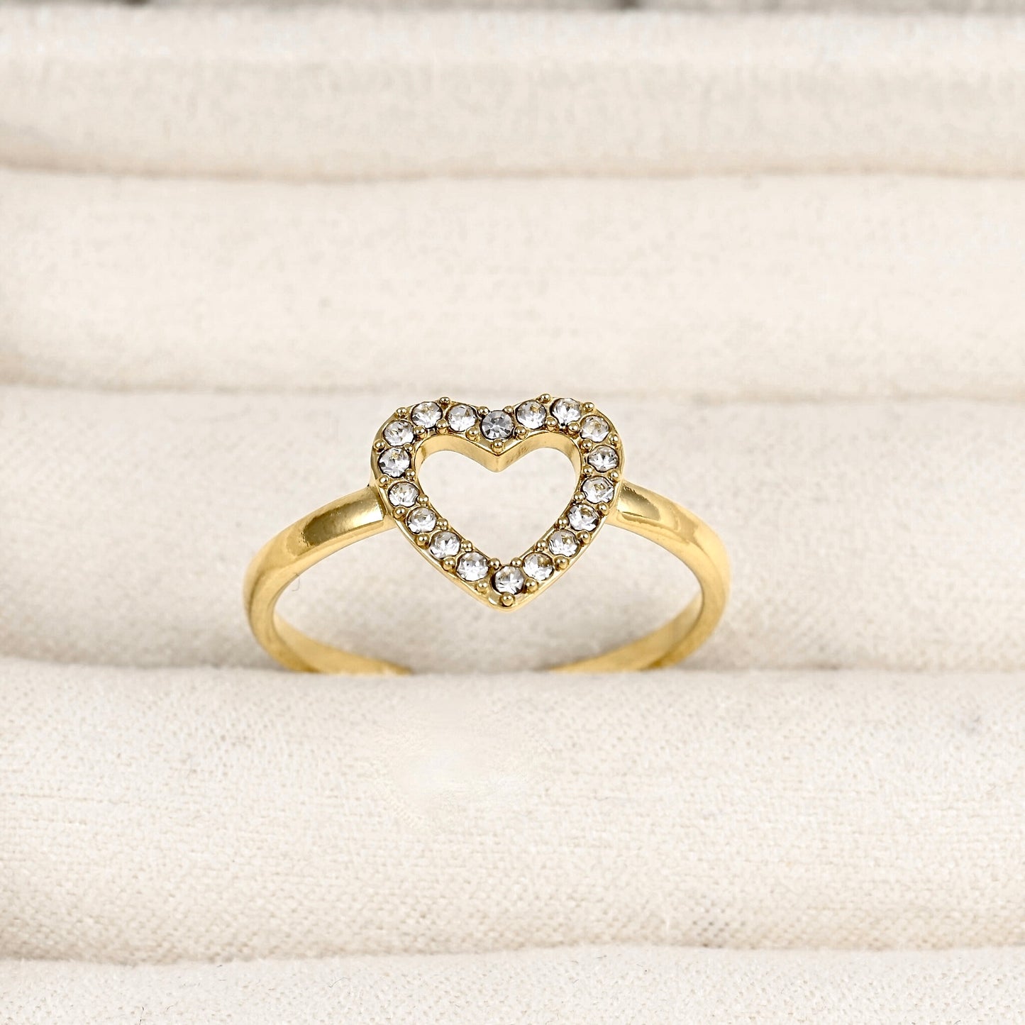 A gold ring featuring an open heart-shaped top set with small clear stones, displayed on a textured cream fabric surface.