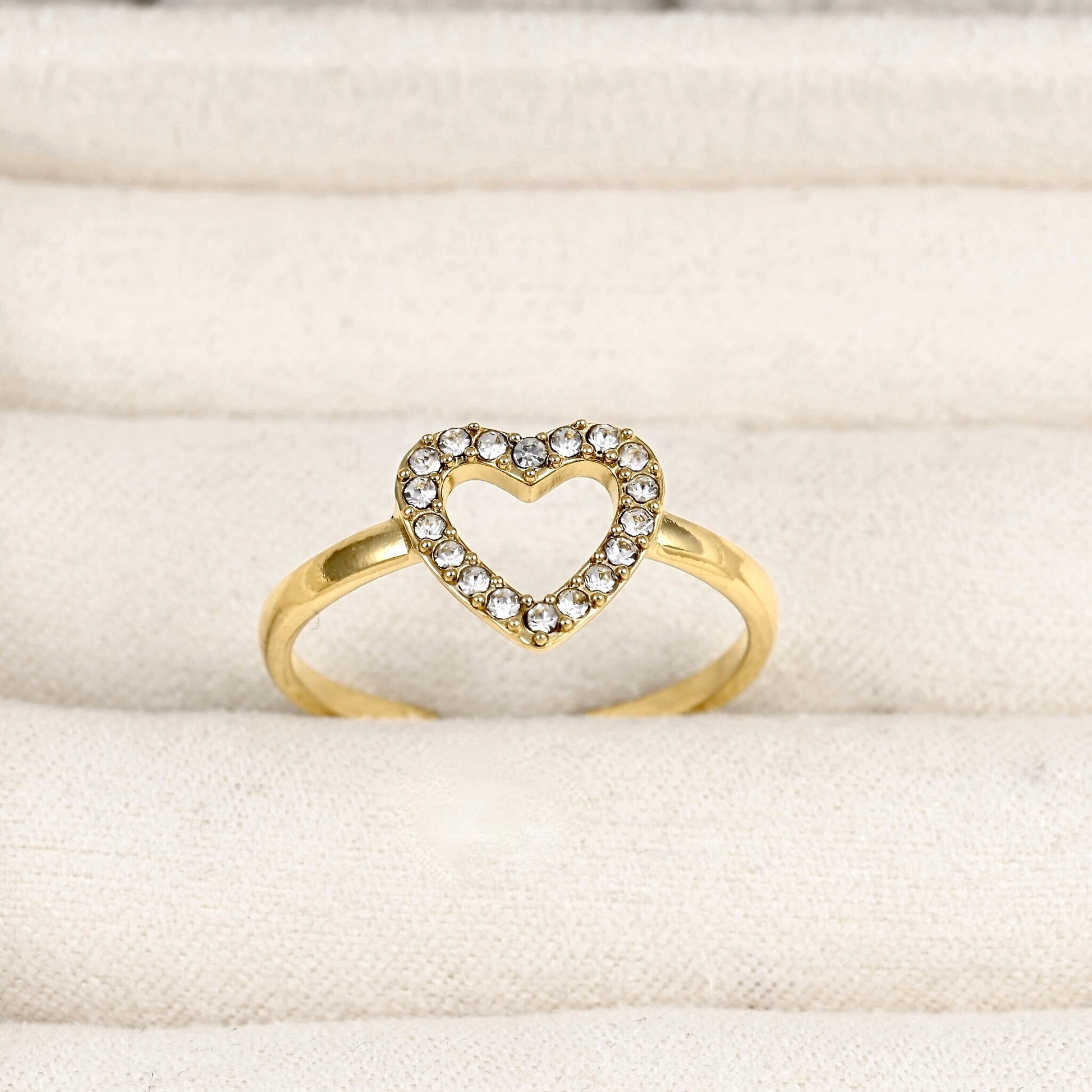 A gold ring featuring an open heart-shaped top set with small clear stones, displayed on a textured cream fabric surface.