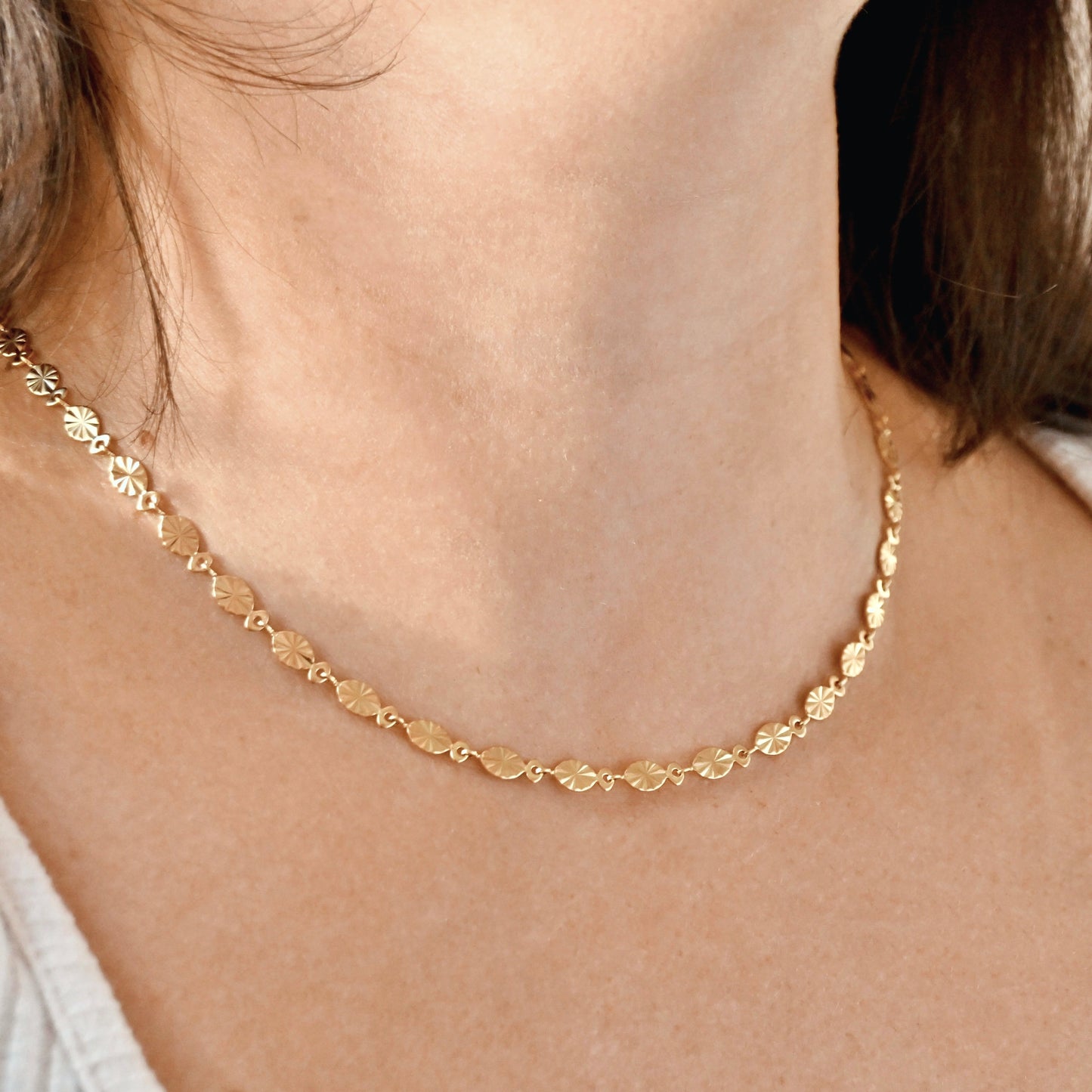 A delicate gold chain necklace with small, flat, leaf-like oval charms, shown worn on a person's neck.