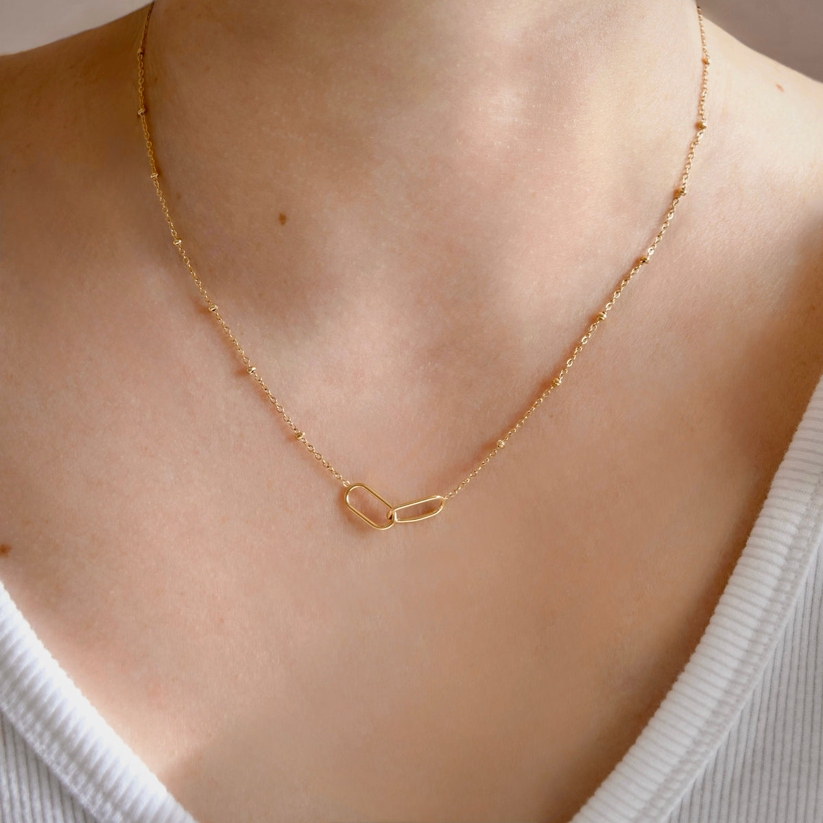 A delicate gold chain necklace with a small open heart pendant, worn on a model's neck over a white ribbed top.