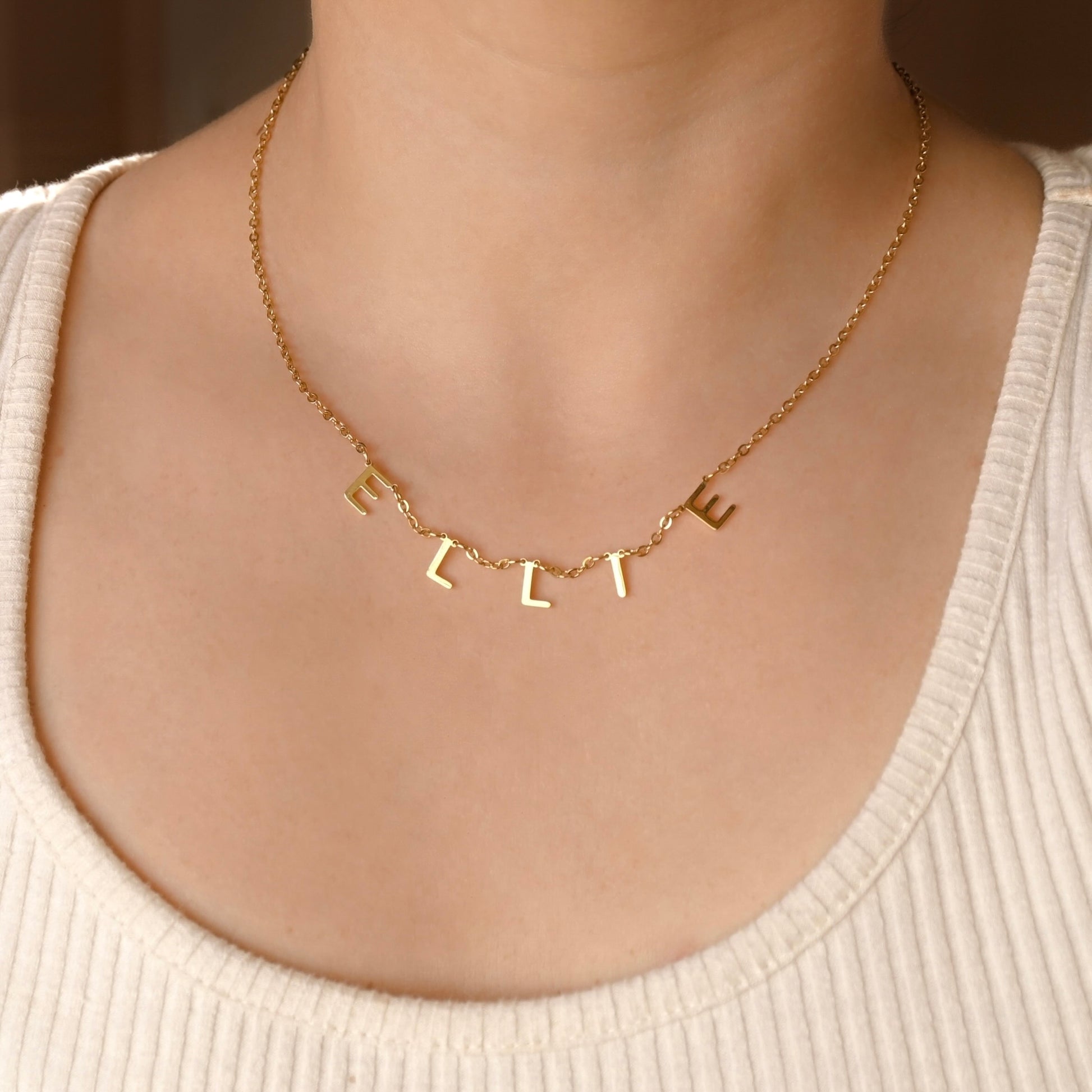 A close-up front view of a person's neck and upper chest wearing a delicate gold chain necklace with individual letter charms spelling 'ELLIE'. The person is wearing a cream-colored ribbed tank top.