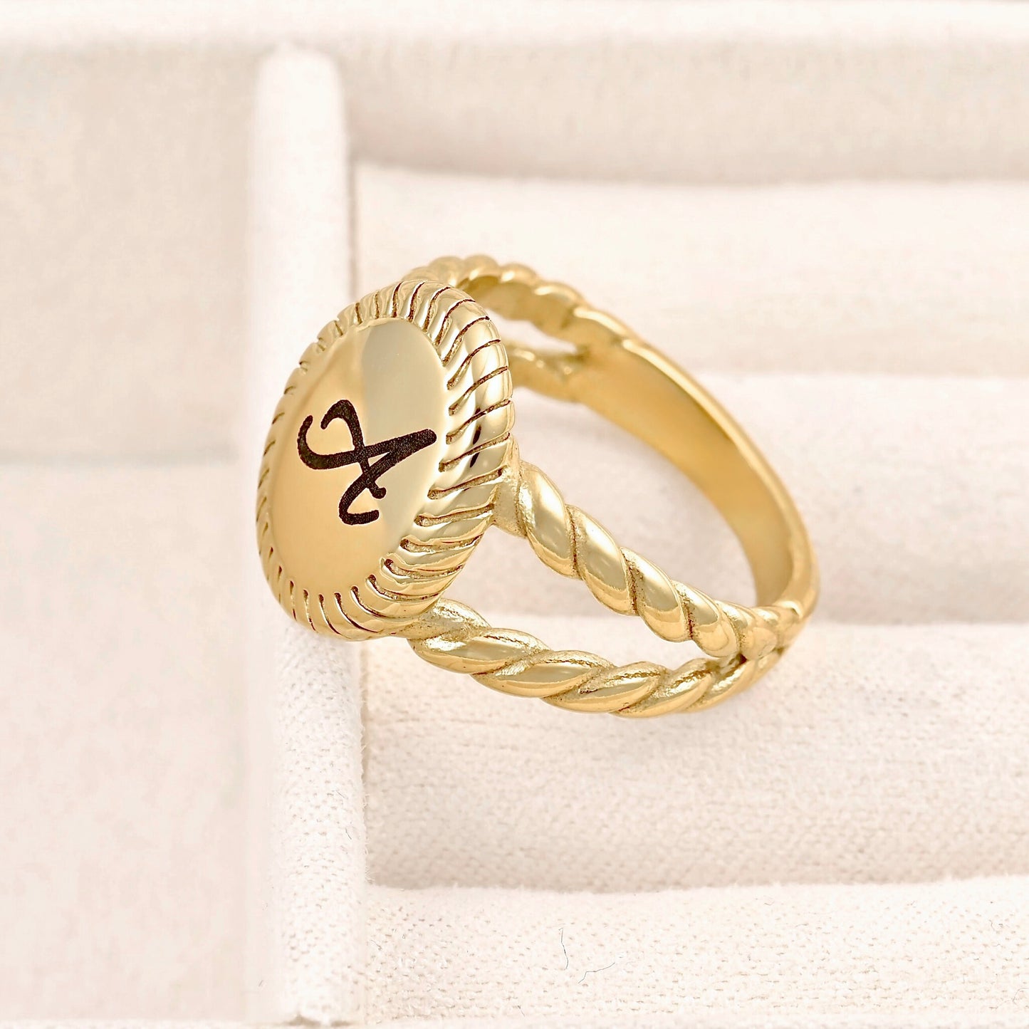 A gold signet ring with a twisted rope band and an oval face engraved with a black monogram, displayed on a cream-colored textured ring box.