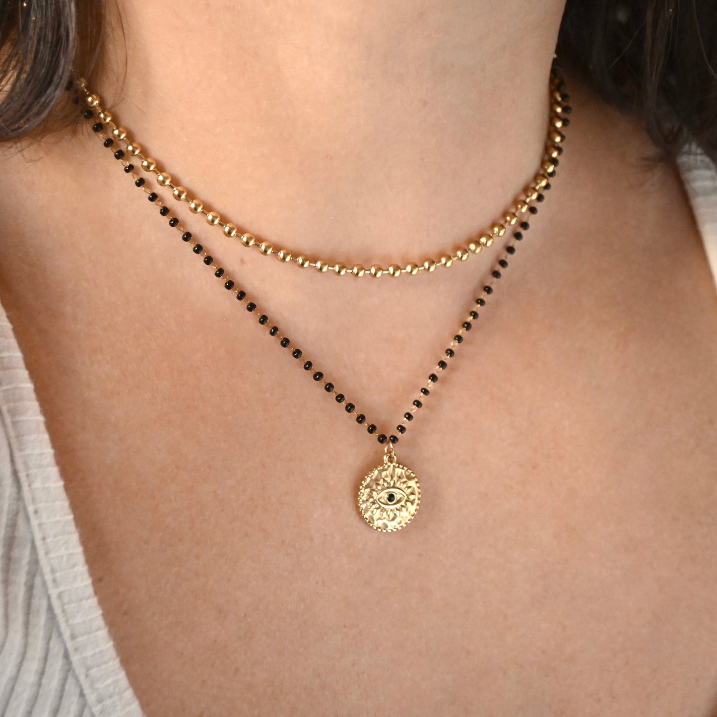 A close-up of a model's neck and chest wearing a double-layered necklace. The top layer is a delicate gold beaded chain, and the bottom layer is a black beaded chain with a textured gold circular pendant featuring an eye design.