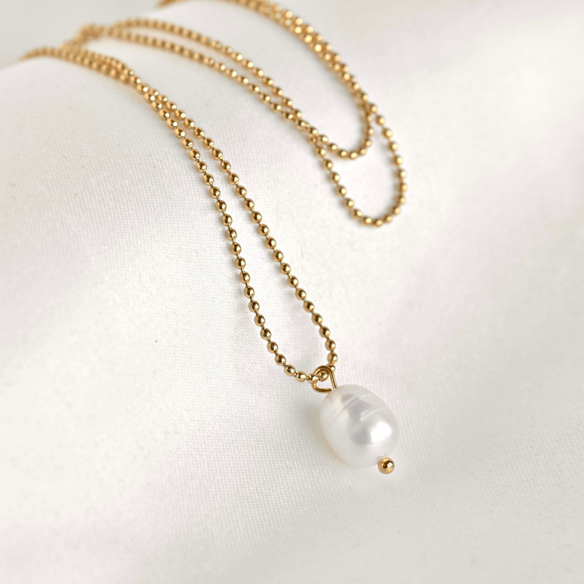 A delicate gold chain necklace with a small white baroque pearl pendant and a tiny gold bead accent, displayed on a soft white fabric background.