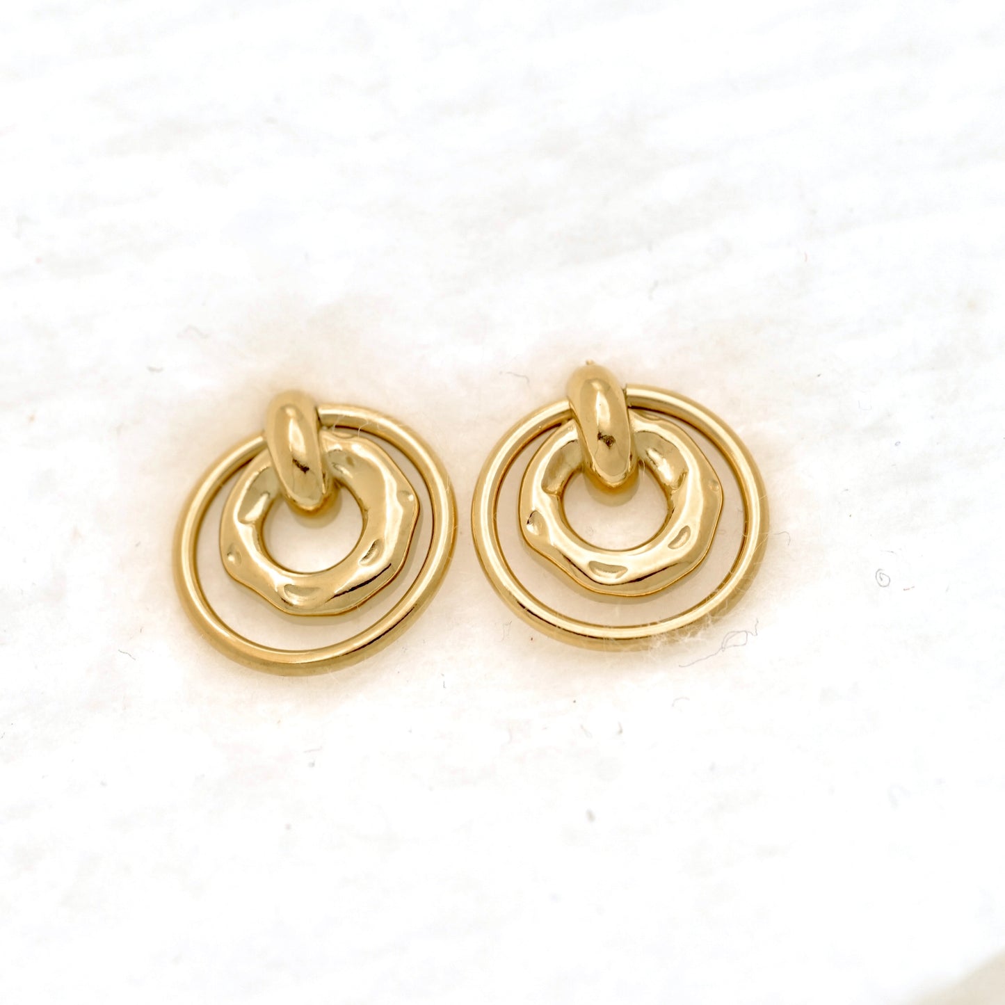 A pair of gold-toned stud earrings with a textured, organic wavy inner ring and a smooth outer circular ring, displayed on a white textured surface.