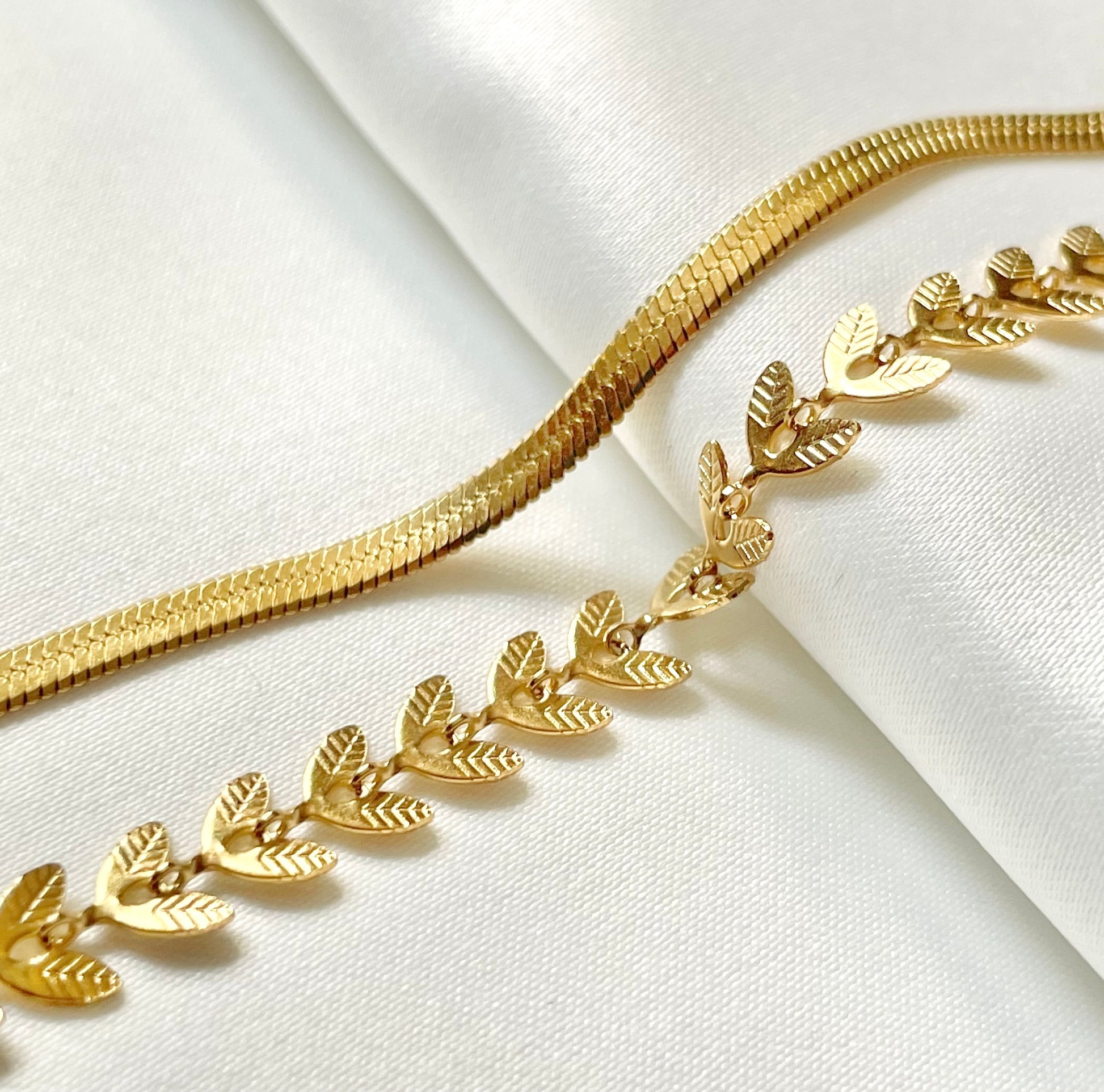 Gold necklace with leaf design and a gold herringbone chain on a white fabric background