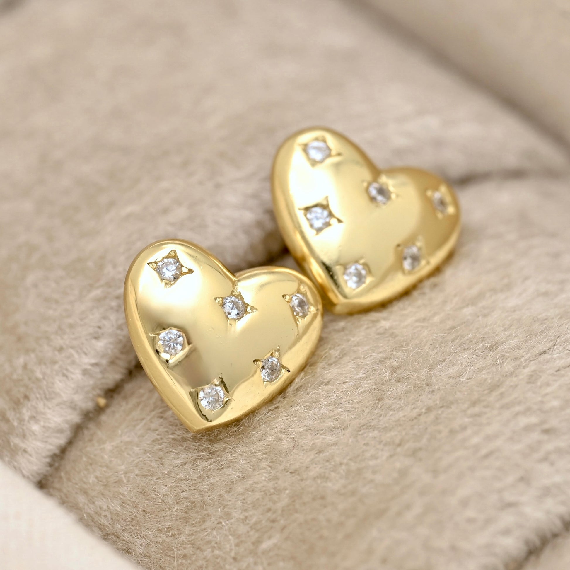 A pair of gold heart-shaped stud earrings, each adorned with small clear gemstones, displayed on a soft beige textured fabric surface.