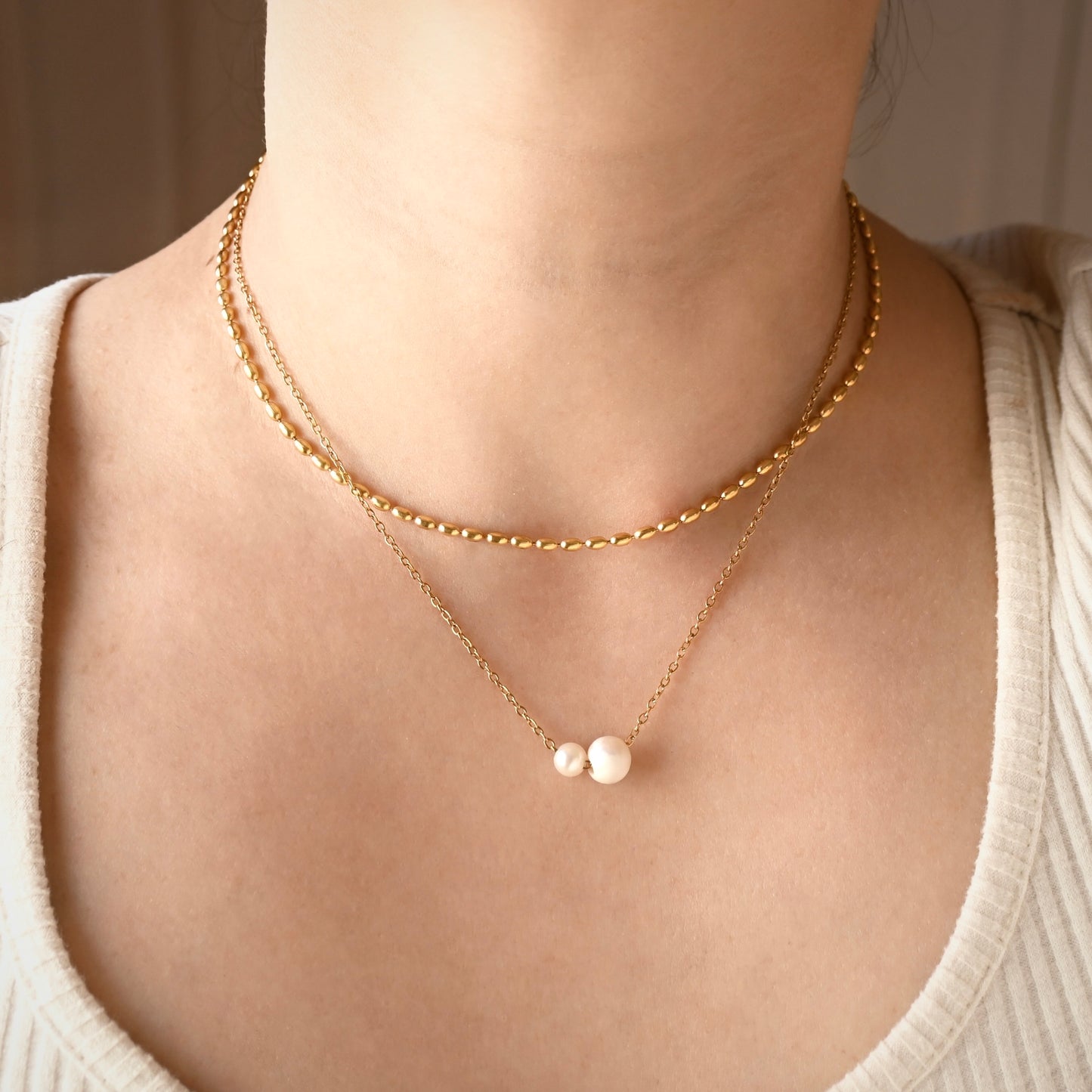 A close-up of a person's neck and upper chest wearing a layered gold necklace set. The set includes a short beaded gold chain and a longer chain with two small white pearl-like beads at the center.