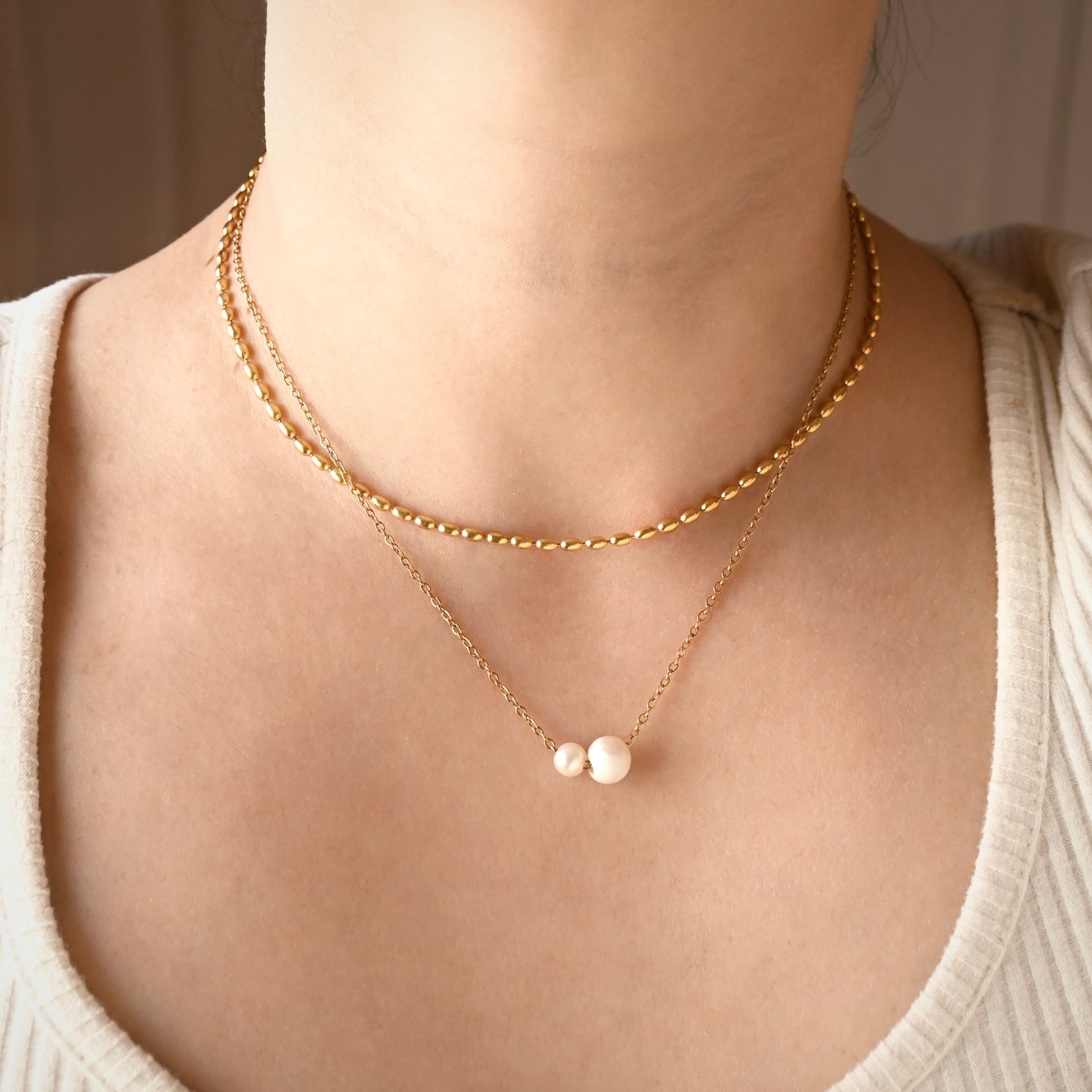 A close-up of a person's neck and upper chest wearing a layered gold necklace set. The set includes a short beaded gold chain and a longer chain with two small white pearl-like beads at the center.