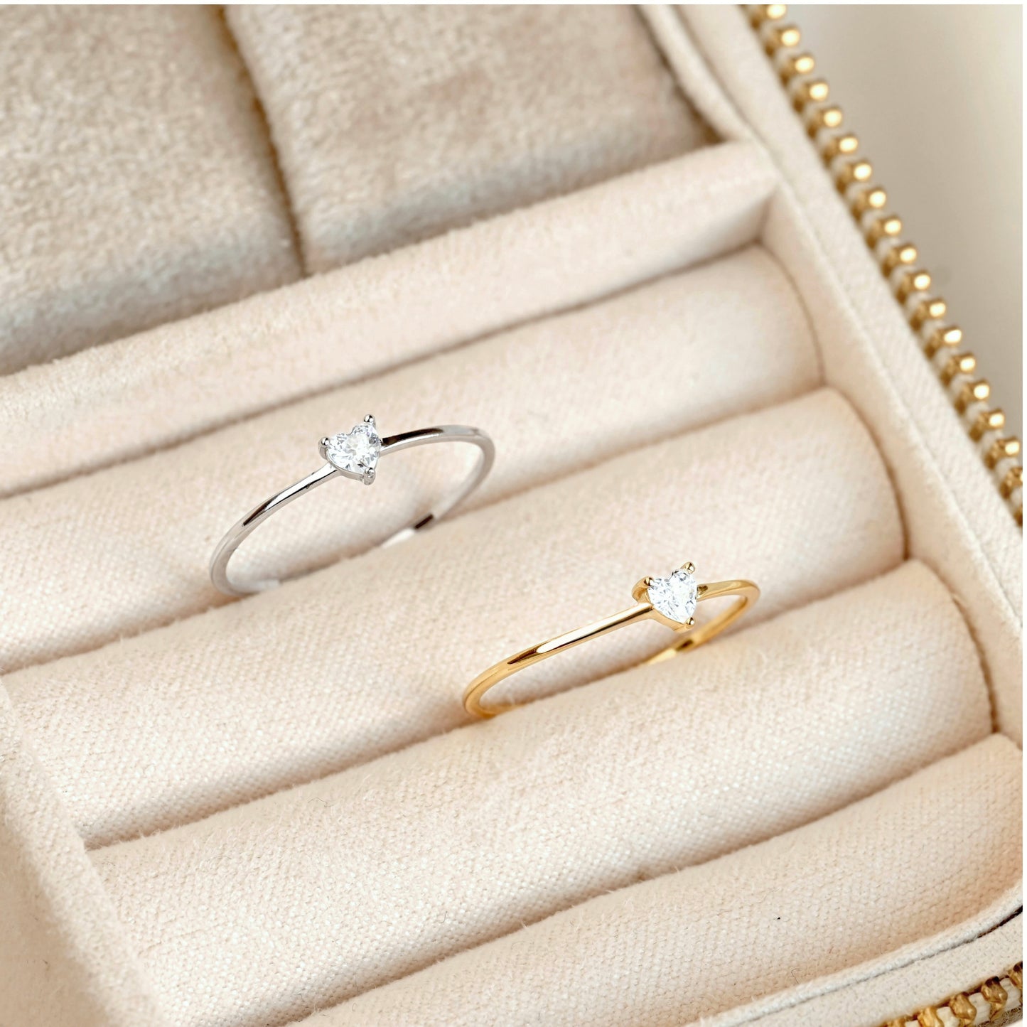 Two slim solitaire rings with heart-shaped clear gemstones, one in silver and one in yellow gold, displayed in a beige velvet ring box.