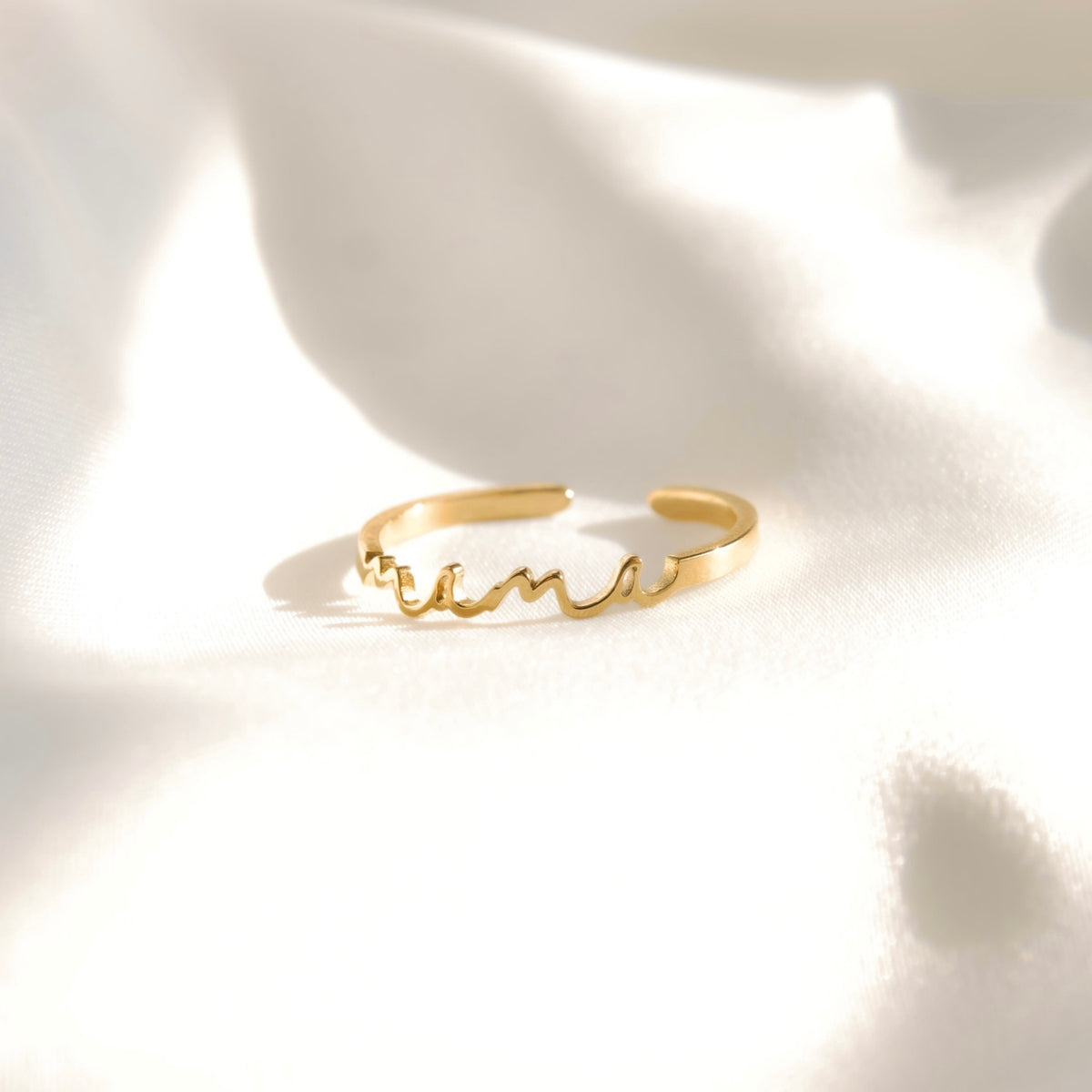 A delicate gold cuff ring with a cursive script design, resting on a softly draped white fabric background with elegant shadows.