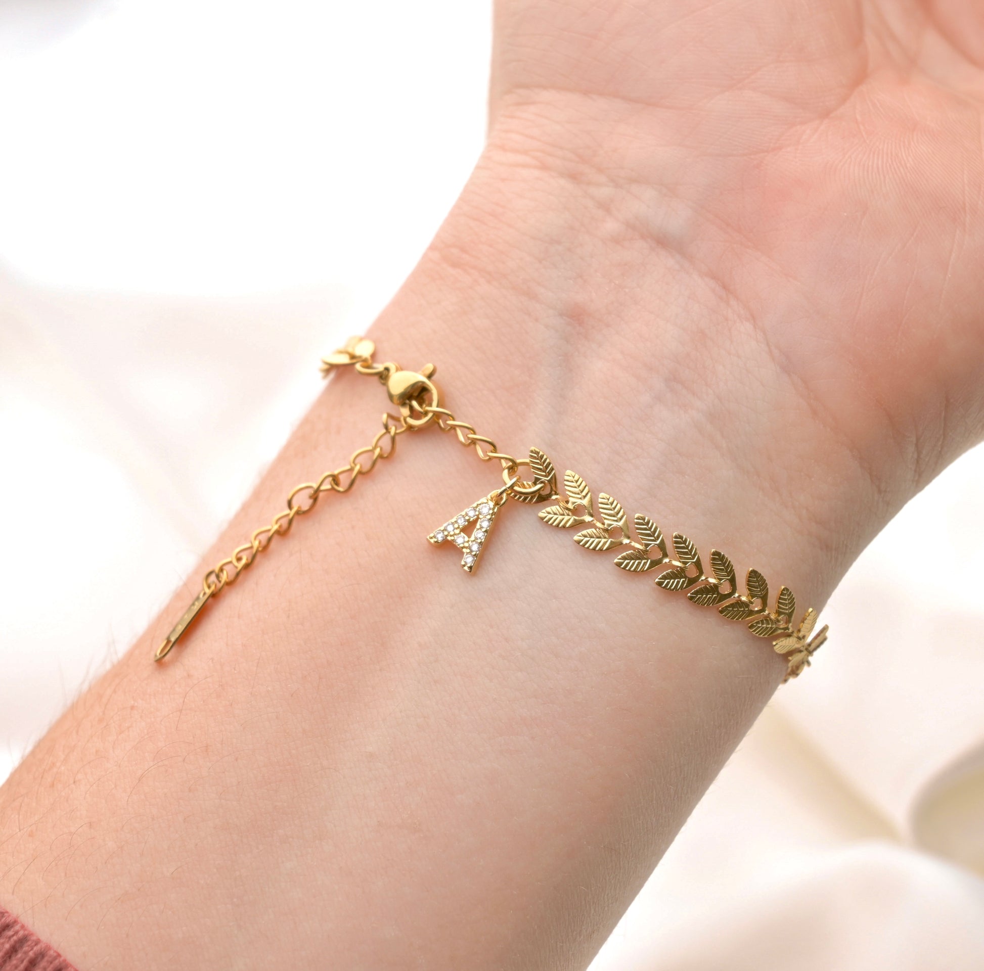 Gold bracelet with leaf links and a diamond 'A' initial charm at the clasp on a wrist against a neutral background