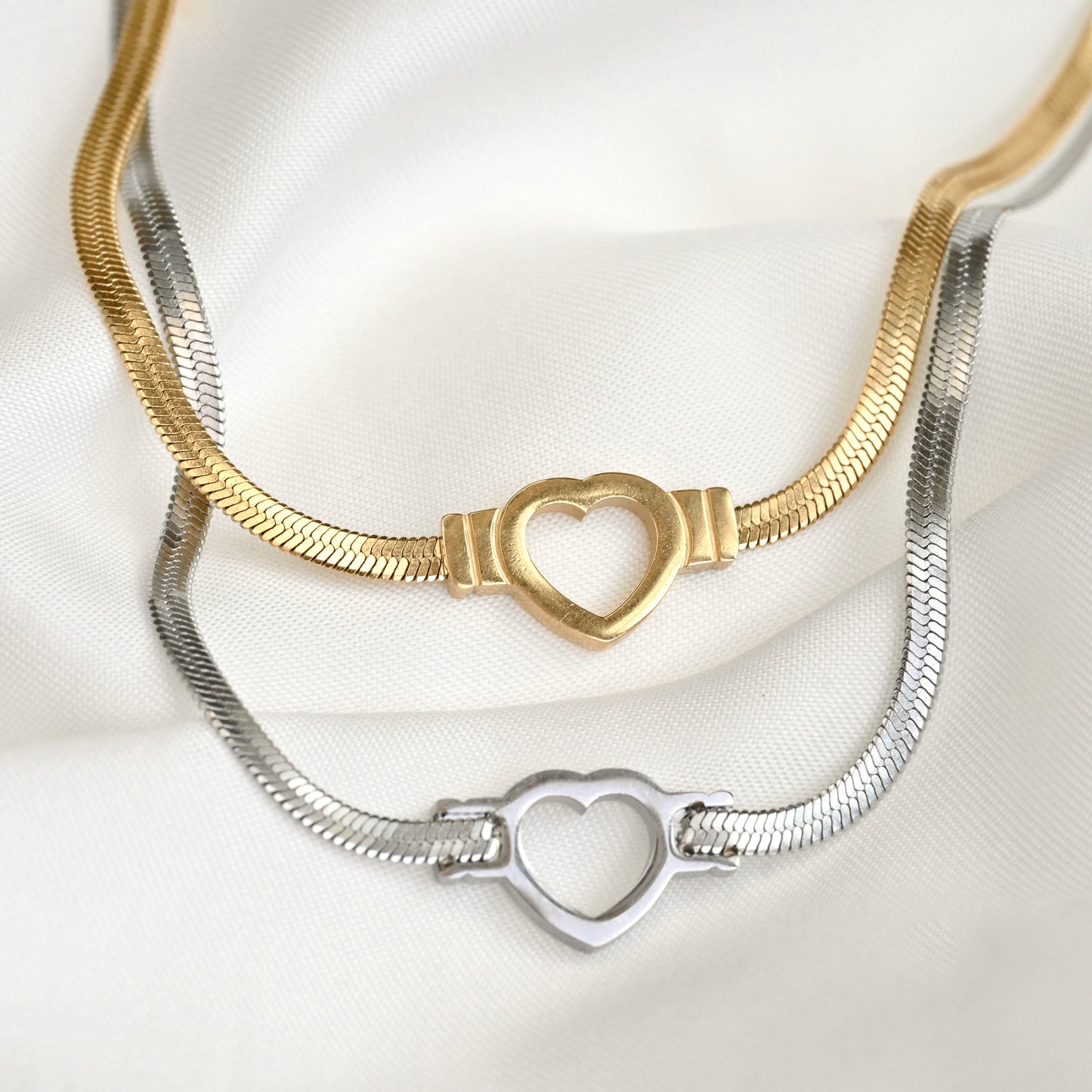 Two snake chain necklaces, one gold and one silver, with heart cut-out charm on a white background.