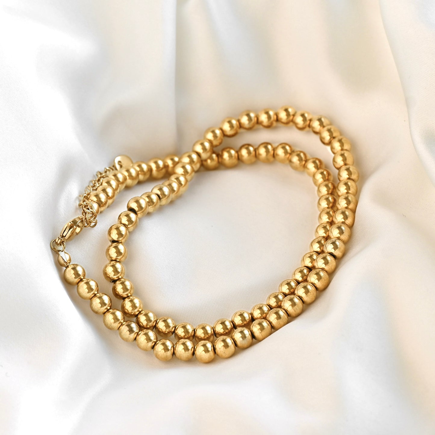 A gold-toned beaded necklace with a matching clasp, arranged in a loop on a draped white satin fabric background.