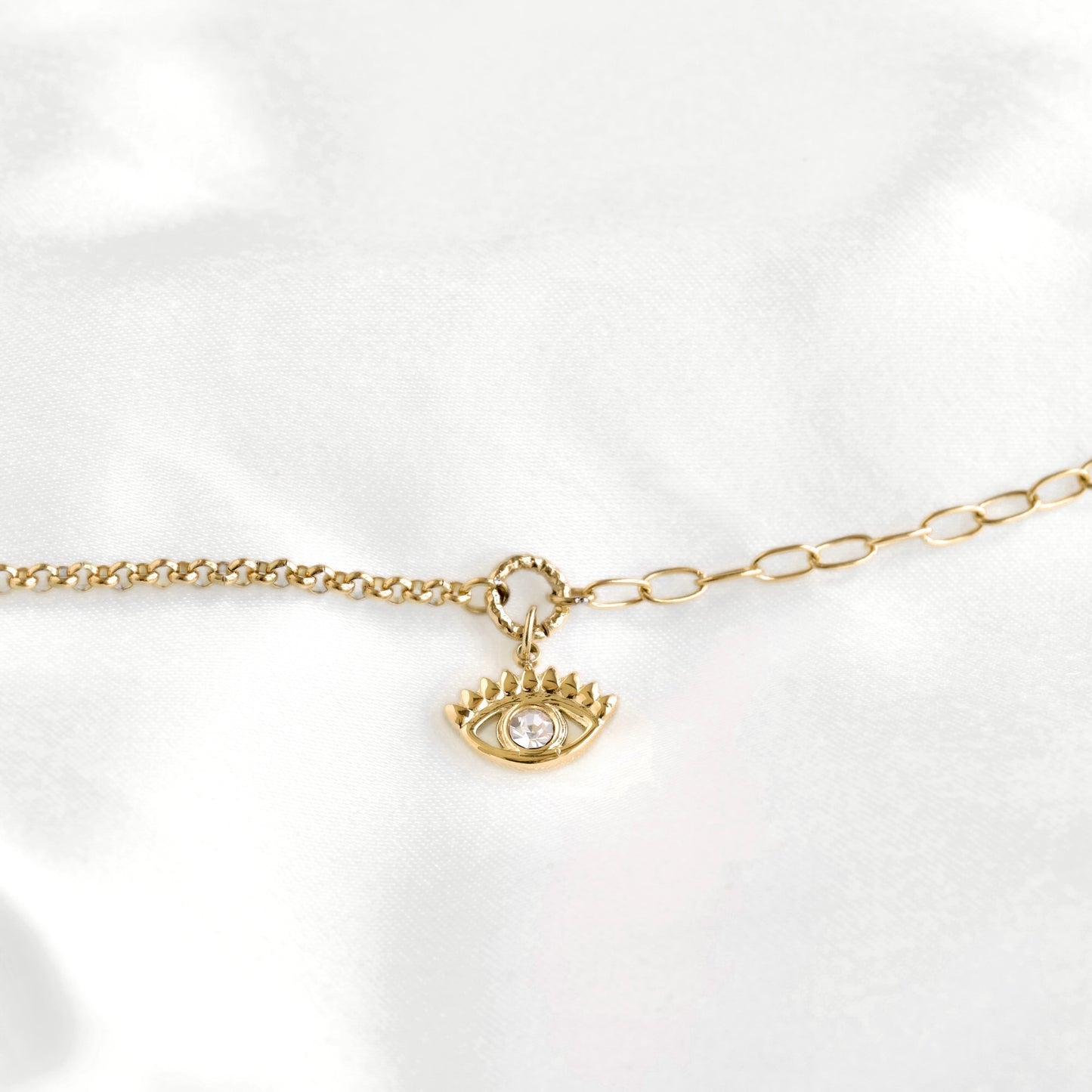 A gold chain bracelet featuring a small gold evil eye pendant with a clear crystal, displayed on a white satin fabric background.