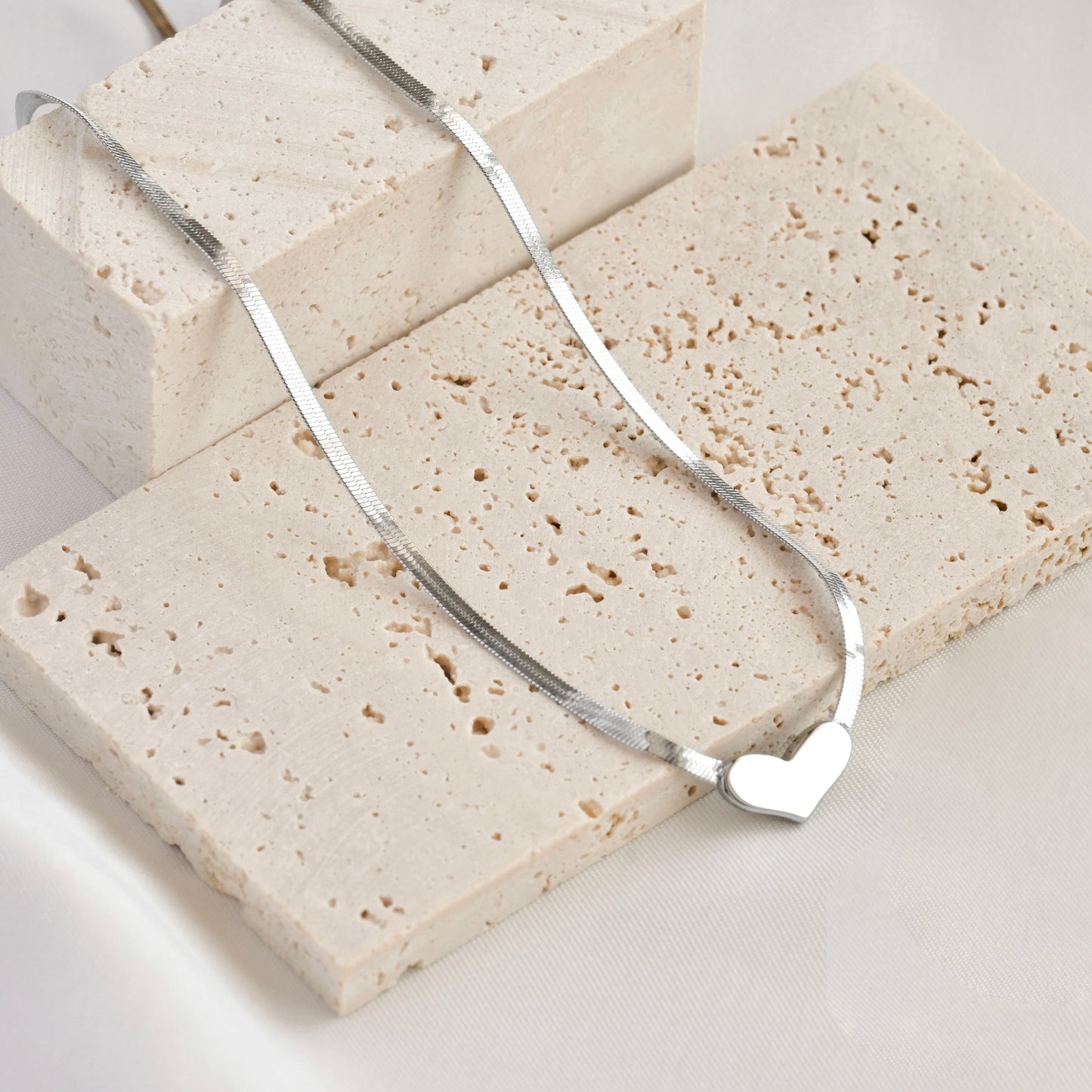 Silver herringbone necklace with a heart pendant on a light stone surface