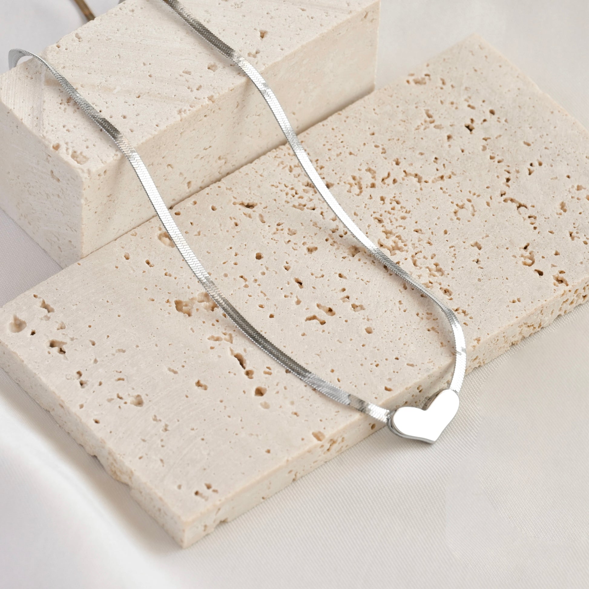 Silver herringbone necklace with a heart pendant on a light stone surface