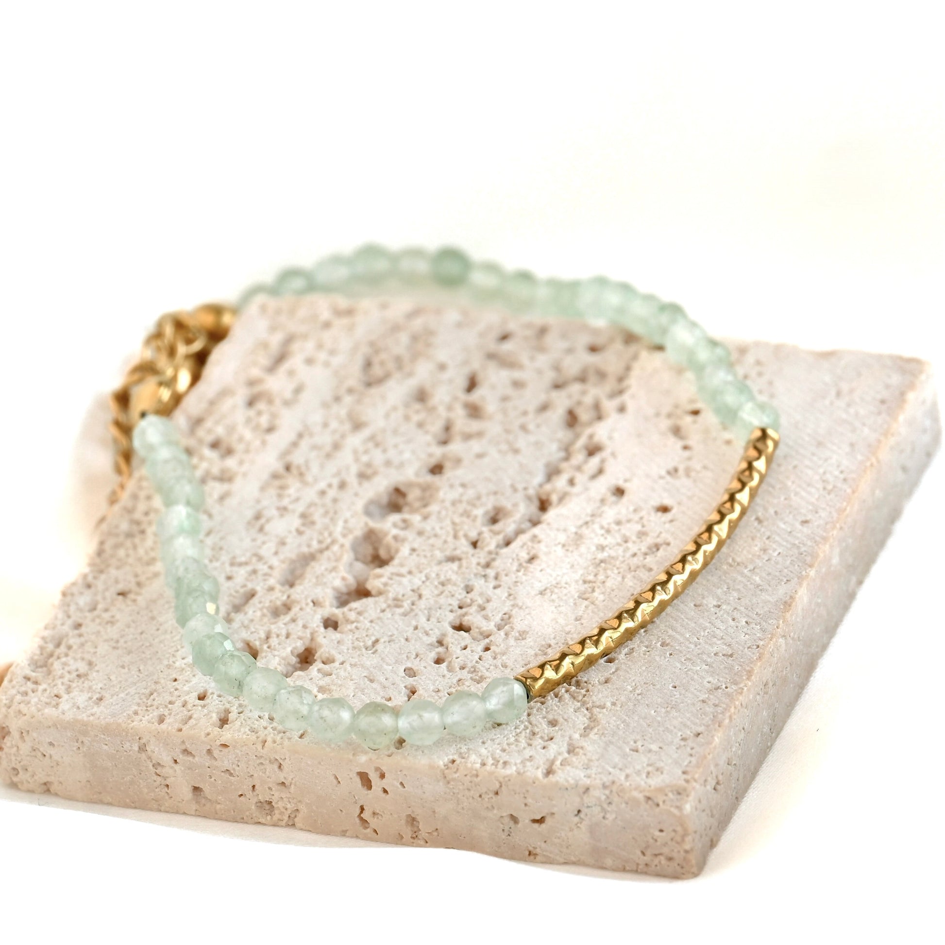 A delicate beaded bracelet with pale mint green beads and a gold chain, displayed on a textured beige stone block against a white background.
