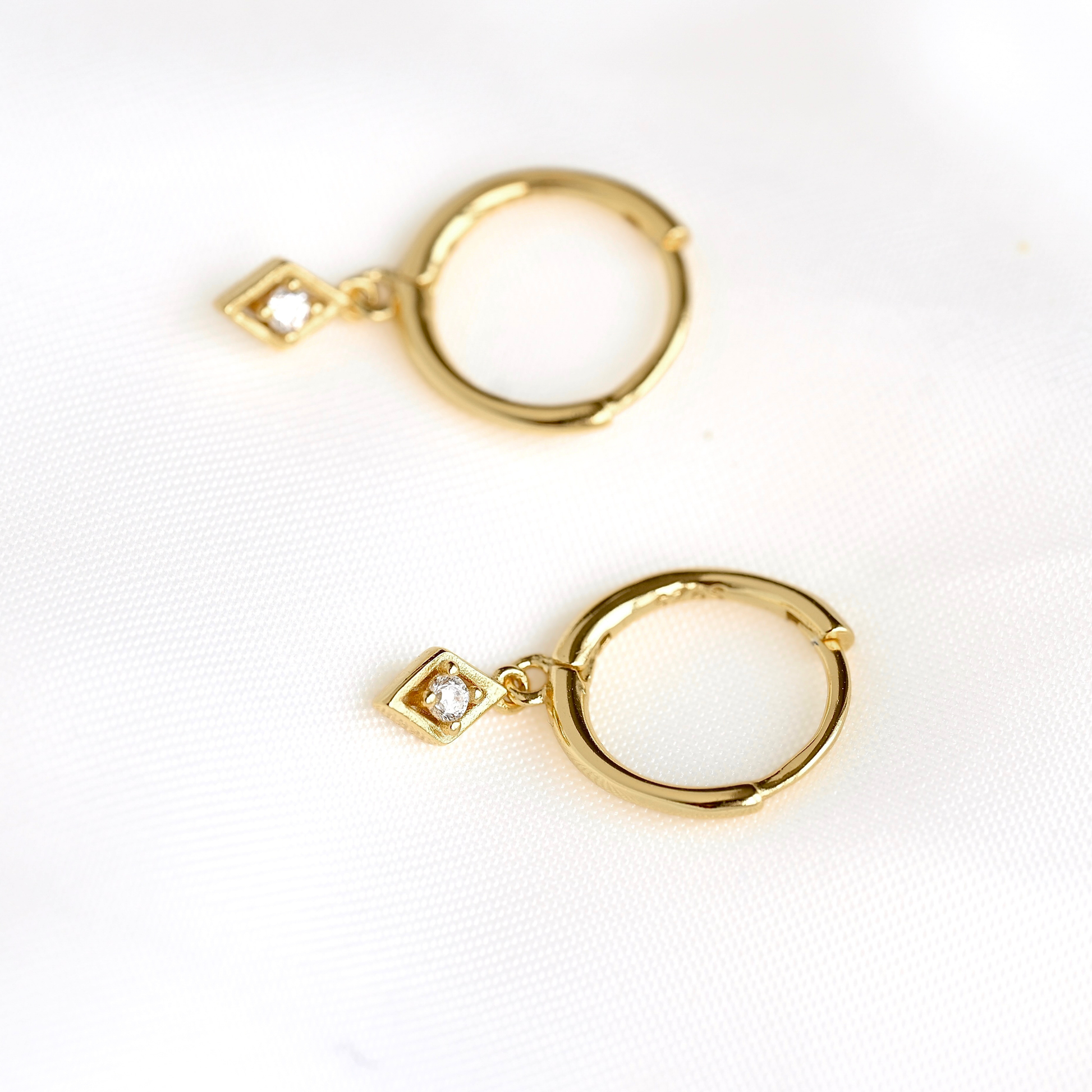 A pair of gold huggie hoop earrings with a small diamond-shaped clear stones, displayed on a white textured fabric background.