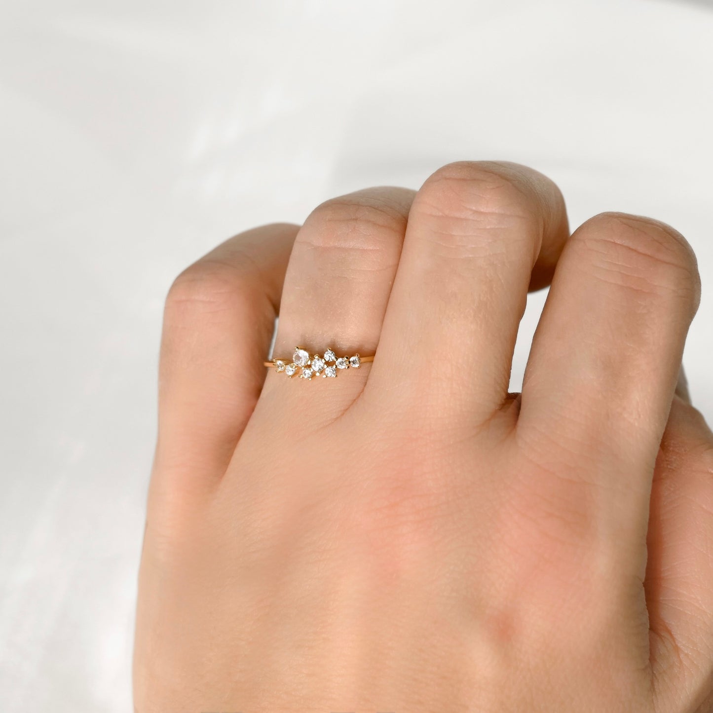 A delicate gold ring with a small cluster of clear gemstones on the middle finger of a hand, photographed against a soft, light gray background.