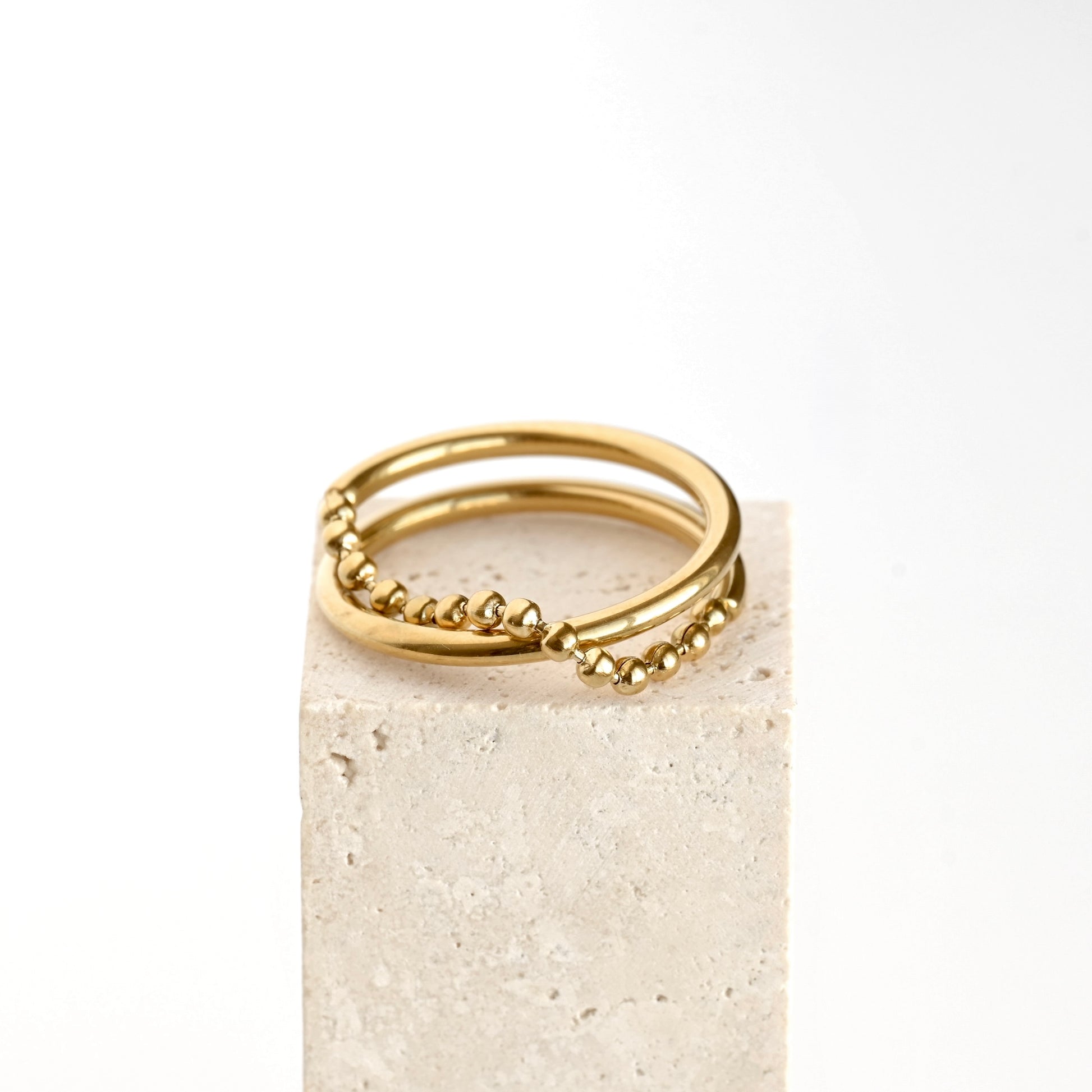 A pair of gold double-band rings with textured beaded detailing, displayed on a light beige stone block against a clean white background.