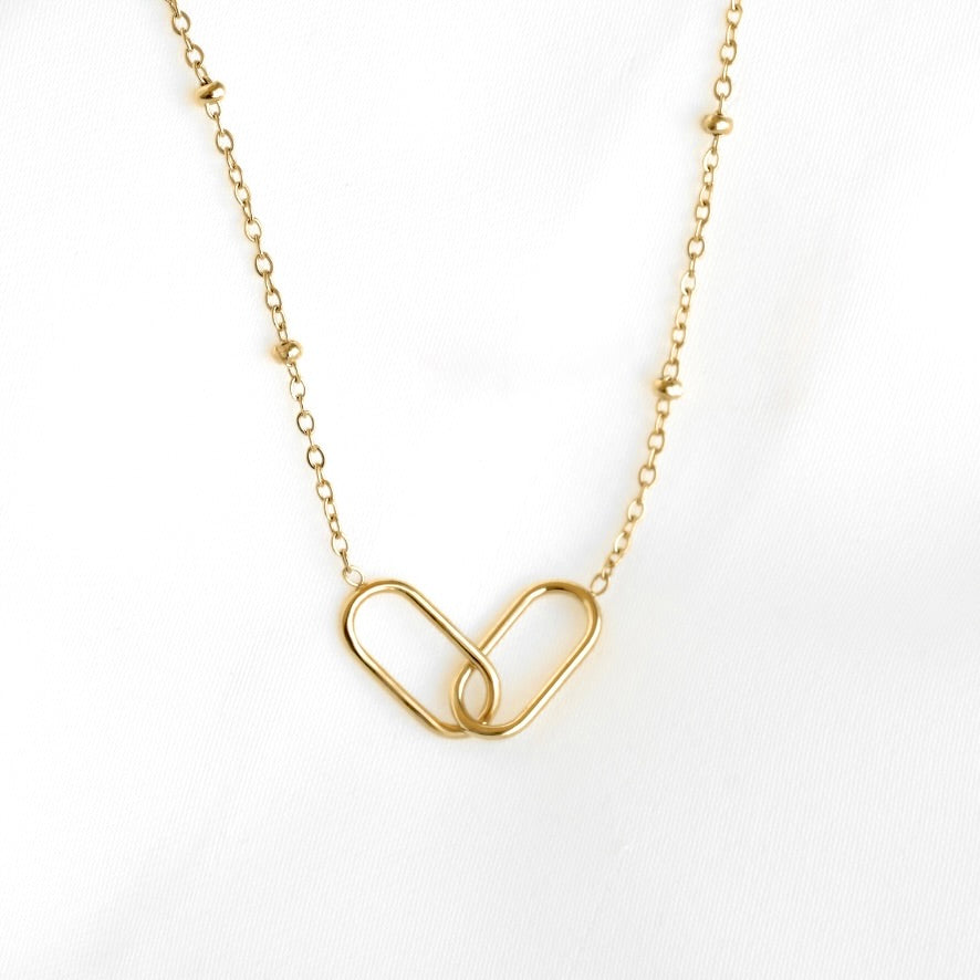 A delicate gold chain necklace featuring a central interlocking double-heart pendant and small bead accents along the chain, displayed on a soft white fabric background.
