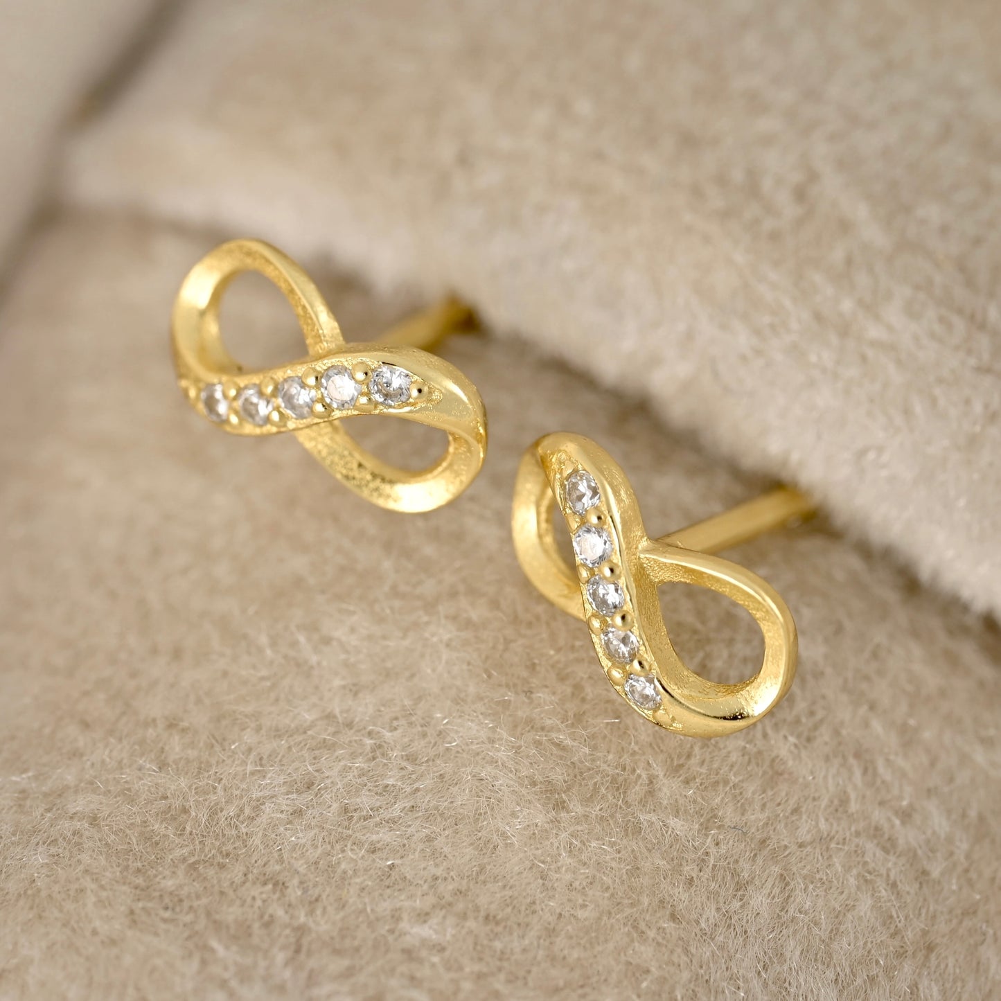 A pair of gold infinity-shaped stud earrings with small clear stones set along the curves, displayed on a soft beige textured surface.