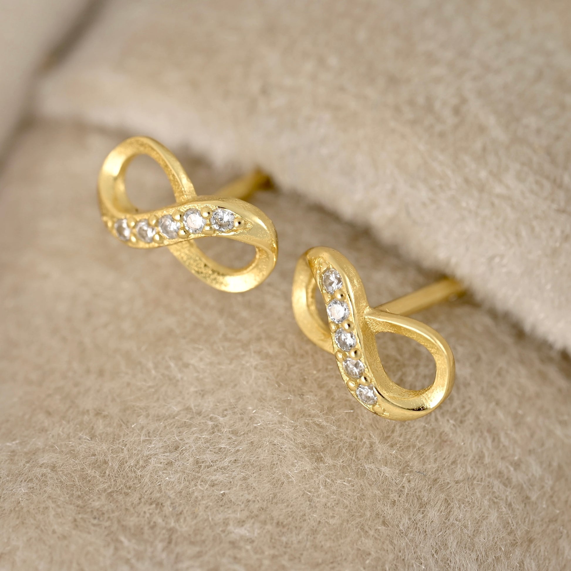 A pair of gold infinity-shaped stud earrings with small clear stones set along the curves, displayed on a soft beige textured surface.