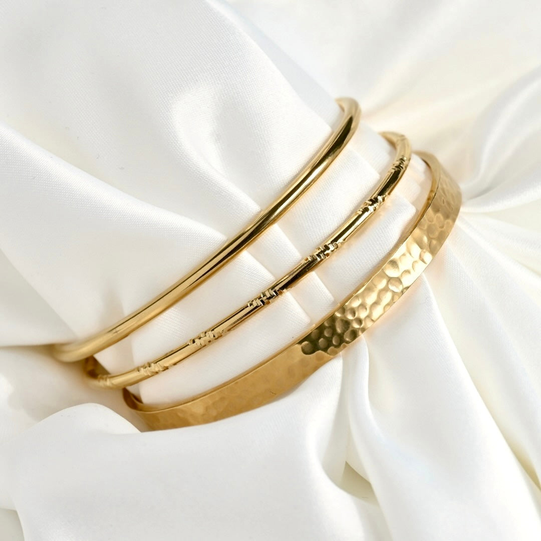 Three gold-toned bangle bracelets displayed on draped white satin fabric, including a smooth polished bangle, a textured beaded bangle, and a hammered bangle.