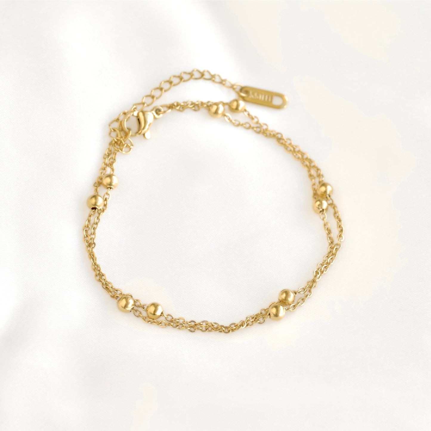 A delicate gold chain anklet with small polished bead accents, displayed on a soft white fabric background.