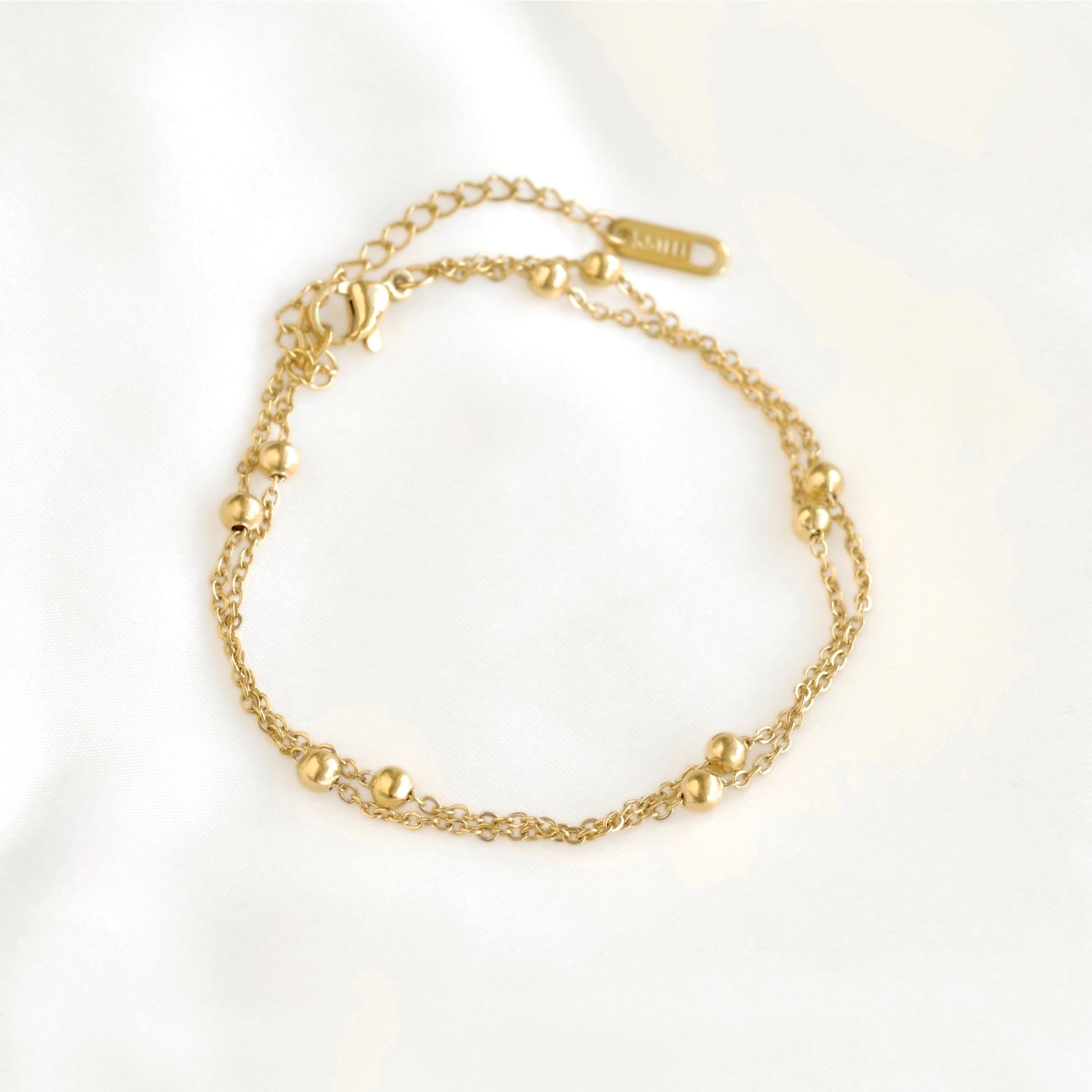 A delicate gold chain anklet with small polished bead accents, displayed on a soft white fabric background.