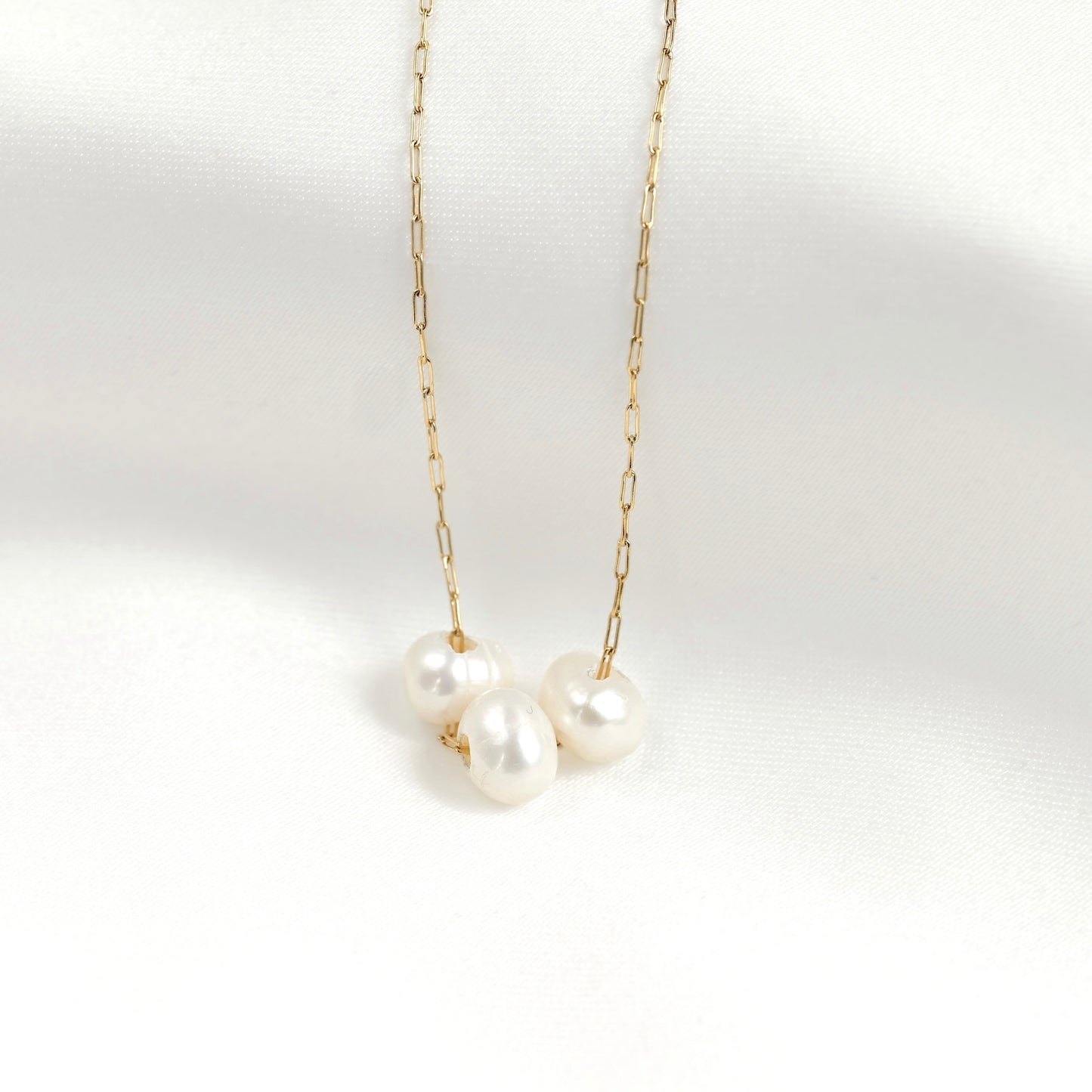 A delicate gold chain necklace featuring three small, lustrous white freshwater pearls clustered at the center, displayed against a clean white fabric background.