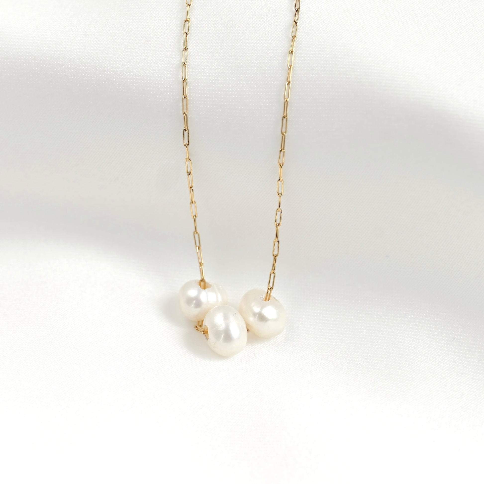 A delicate gold chain necklace featuring three small, lustrous white freshwater pearls clustered at the center, displayed against a clean white fabric background.