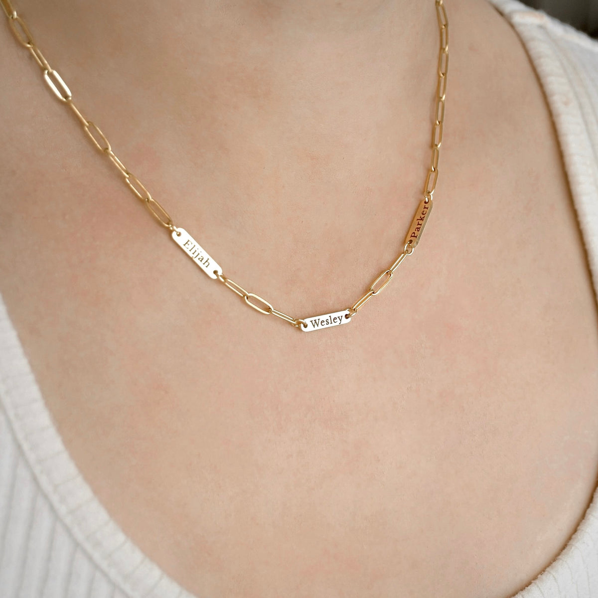 A close-up of a gold chain necklace worn on a person's neck, featuring three personalized name plates: 'Elijah', 'Wesley', and 'Grace'.