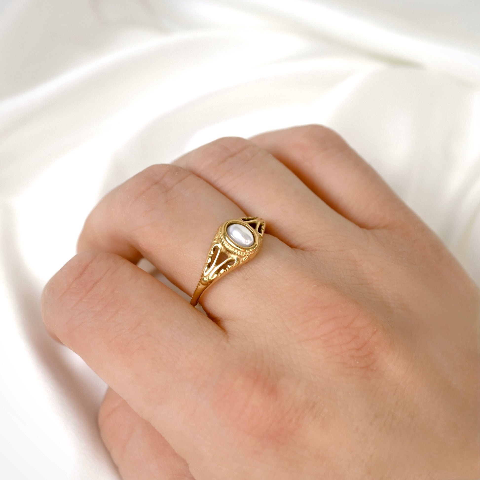 A vintage style gold ring with an oval pearl, worn on a person's finger against a soft white satin background.