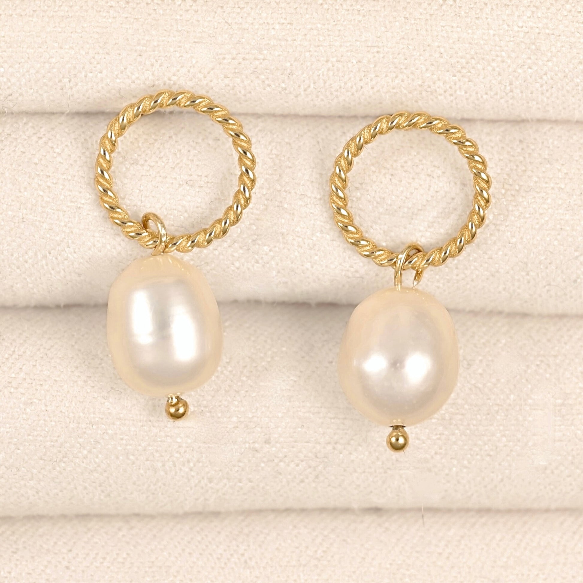 A pair of gold hoop earrings with large, irregular white freshwater pearls hanging from twisted rope-style hoops, displayed on a textured cream fabric background.