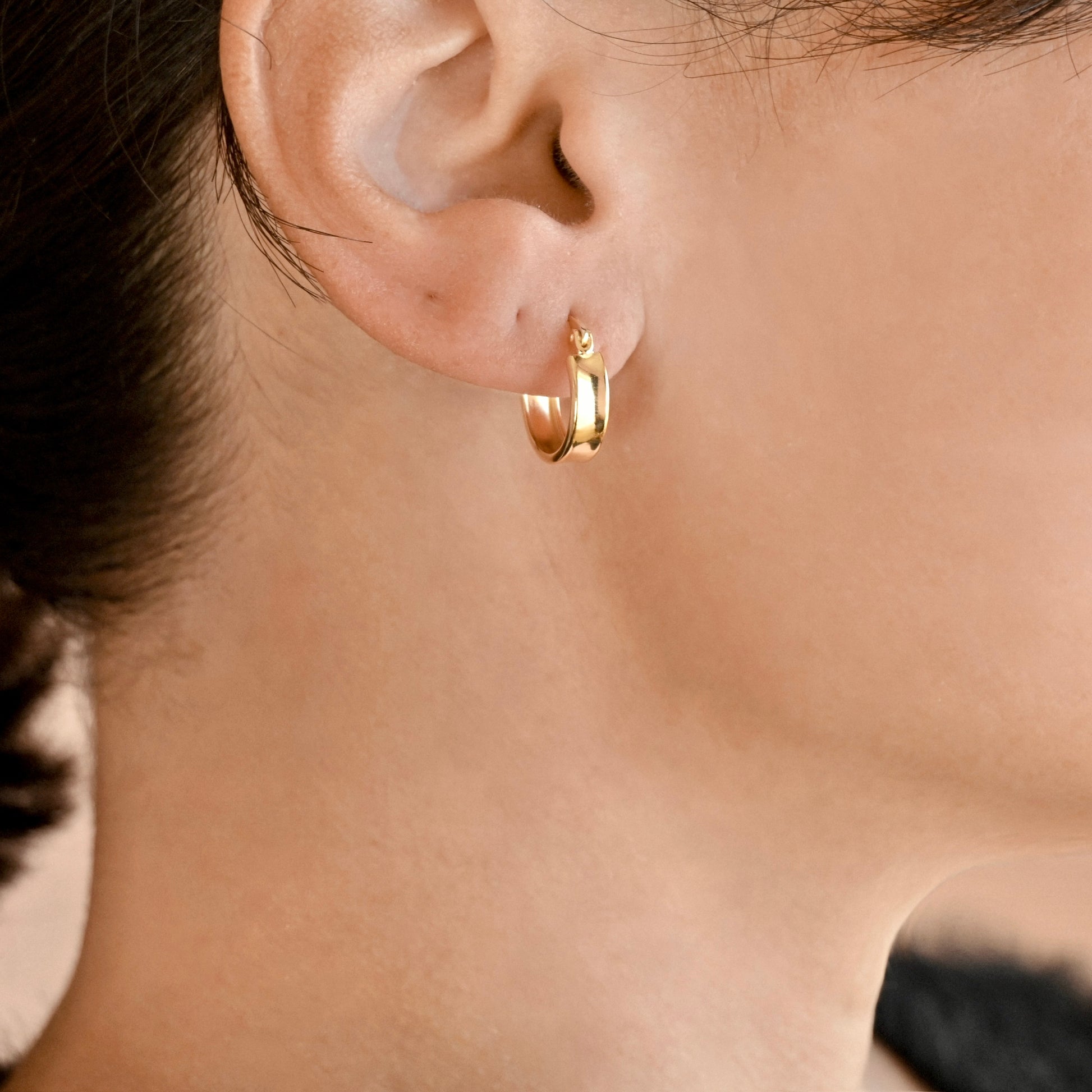A close-up of a person's ear wearing a small polished wide gold hoop earring.