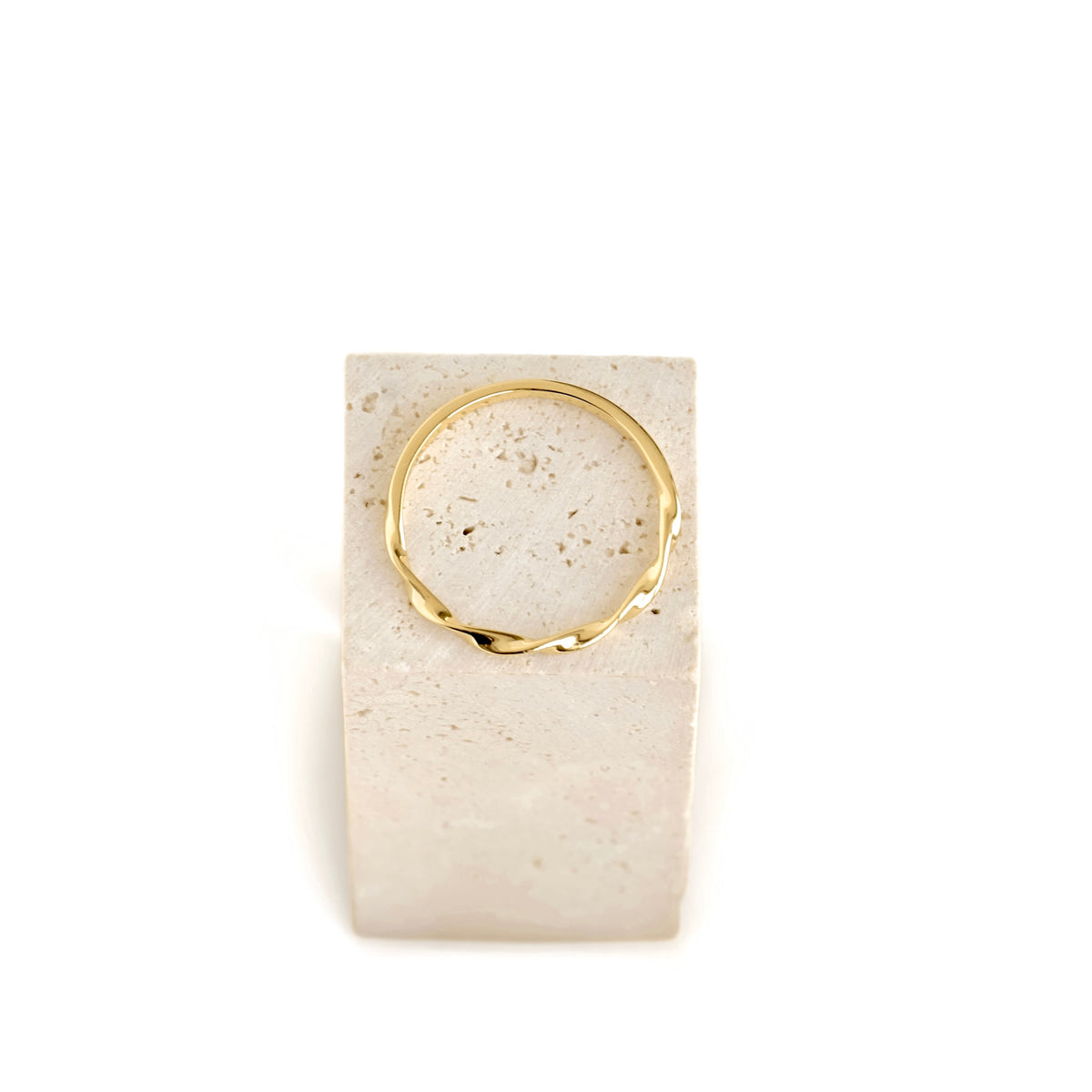 A thin, polished gold ring with a wavy, organic twisted design, displayed on a rectangular beige travertine stone block against a solid white background.