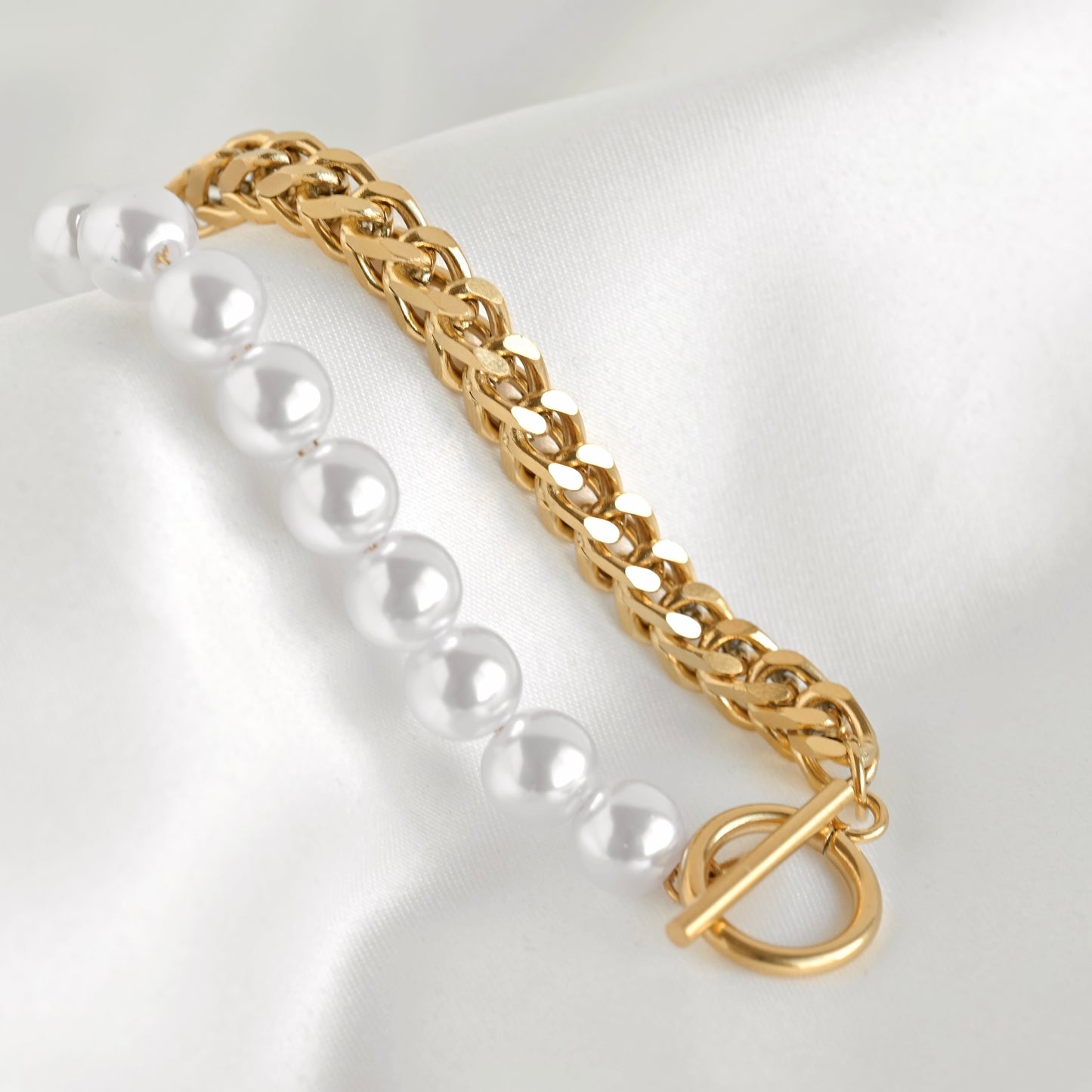 A gold franco chain bracelet with a toggle clasp paired with a strand of white faux pearls, displayed on a white satin fabric background.