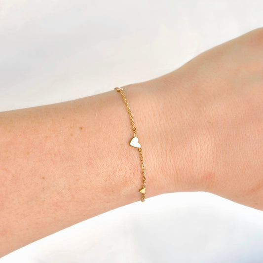 A delicate gold chain bracelet with a small heart charm and two tiny gold heart accents, shown worn on a person's wrist against a soft white background.
