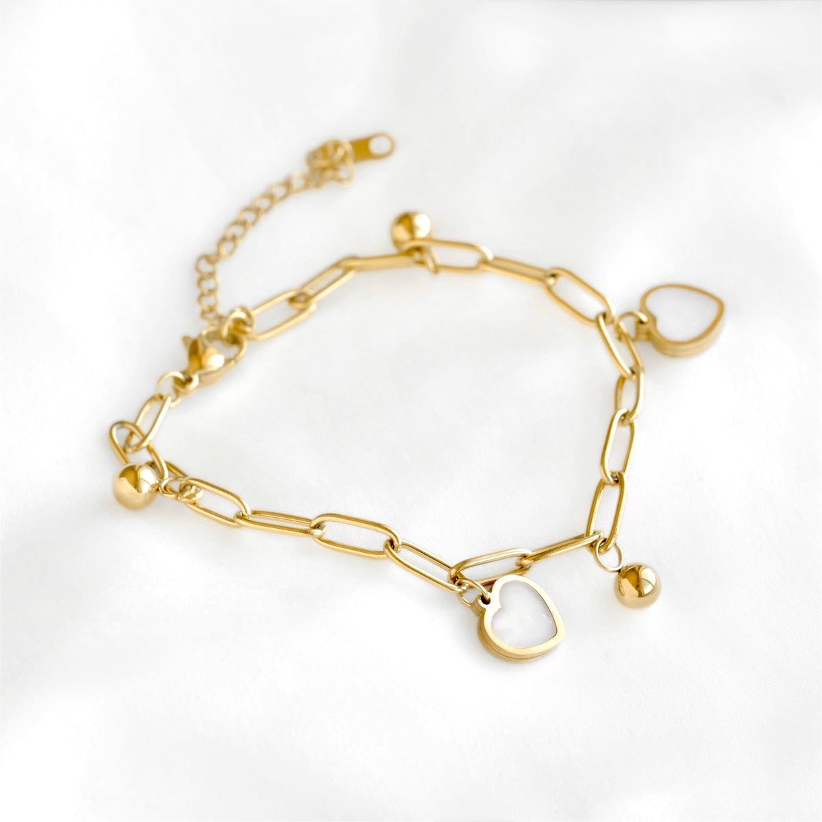 A gold chain bracelet with paperclip links, small bead charms, and two shell-inlay heart-shaped pendants, displayed on a soft white fabric background.