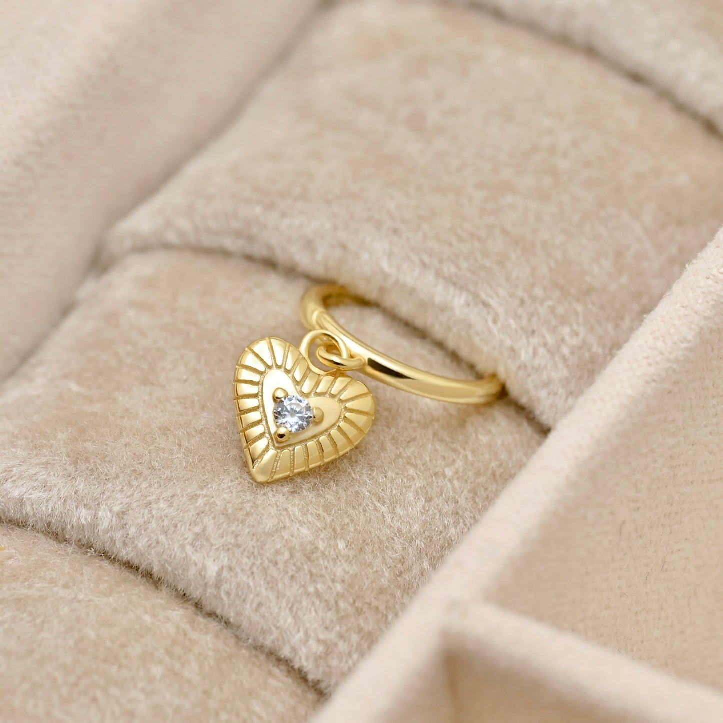 A gold heart-shaped earring with a small clear gemstone in the center, displayed on a beige velvet jewelry box.