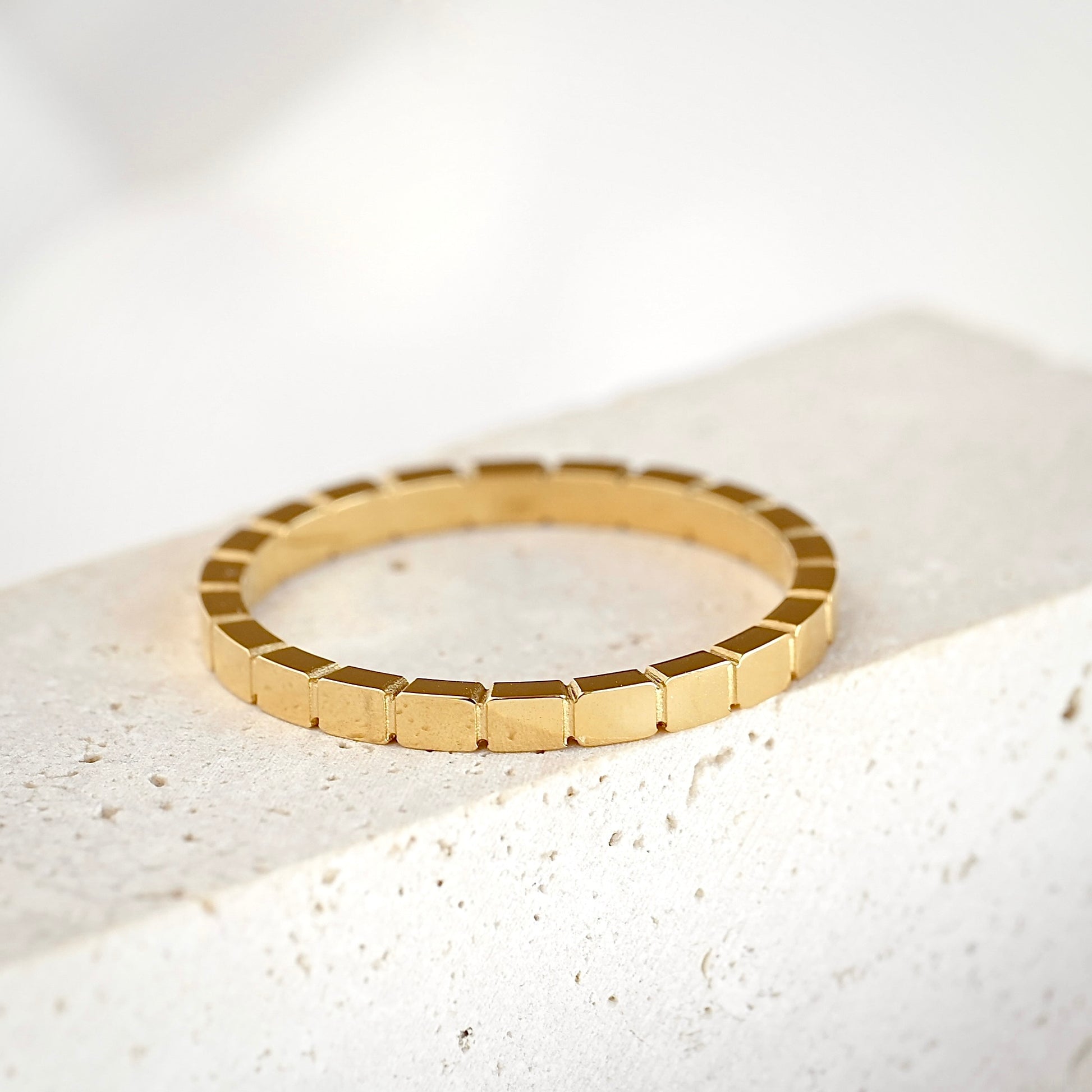 A gold segmented bangle bracelet with a textured, geometric block design, resting on a light beige stone surface.