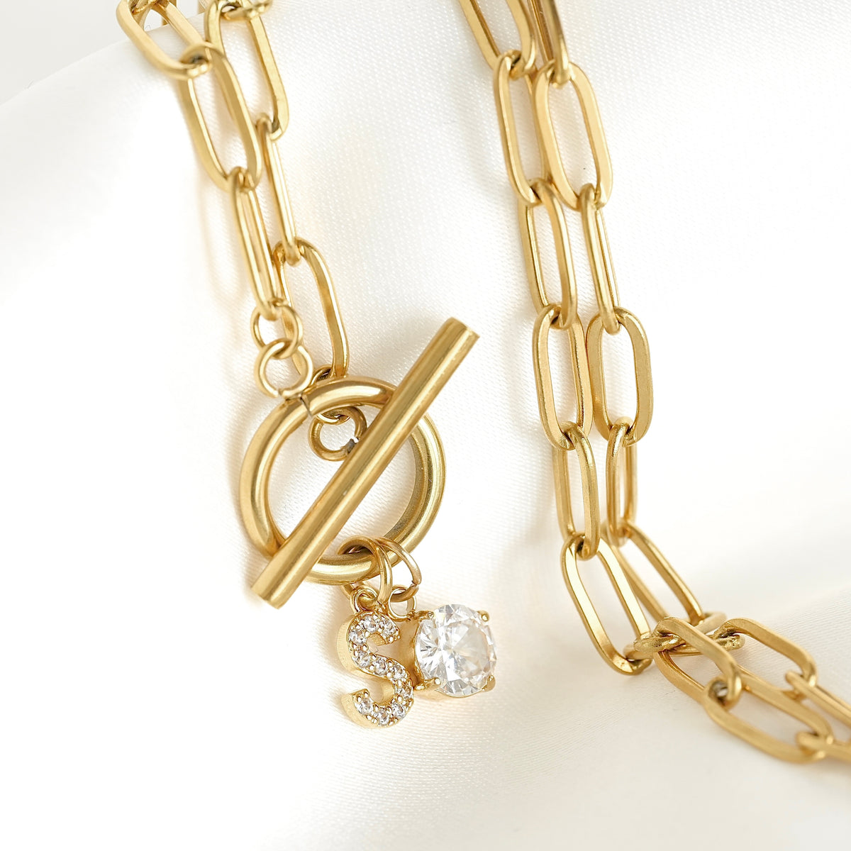 Gold chain necklace with a toggle clasp and a dangling 'S' charm embellished with small clear stones, displayed on a white fabric background.